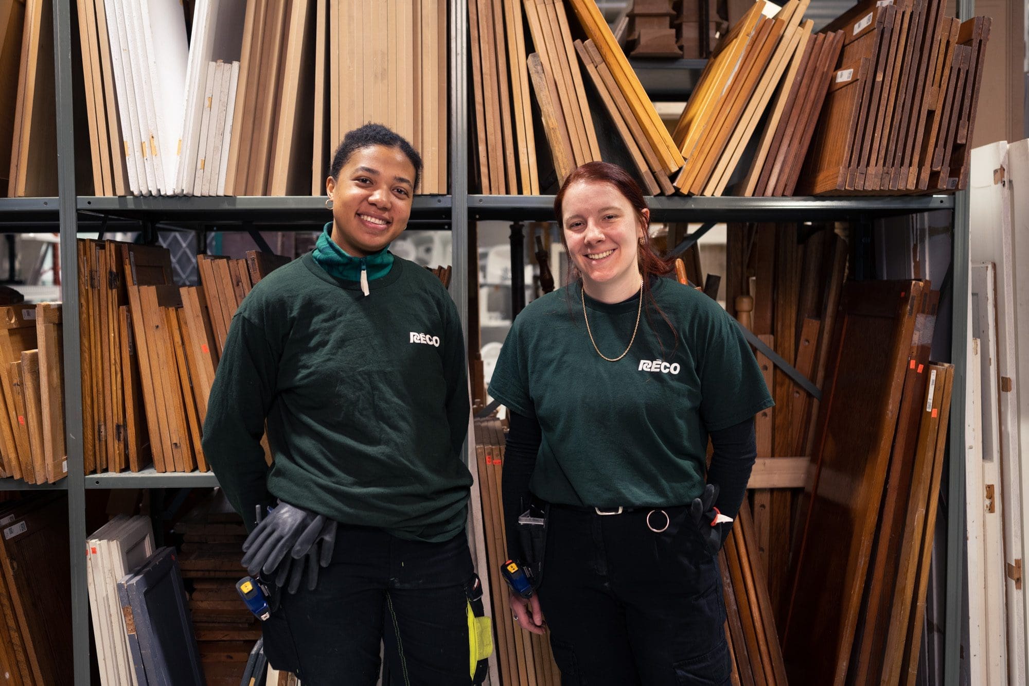 "Deux personnes portant des chemises vertes RECCO se tiennent en souriant devant des étagères remplies de diverses planches de bois. Dans ce qui semble être un atelier ou une zone de stockage