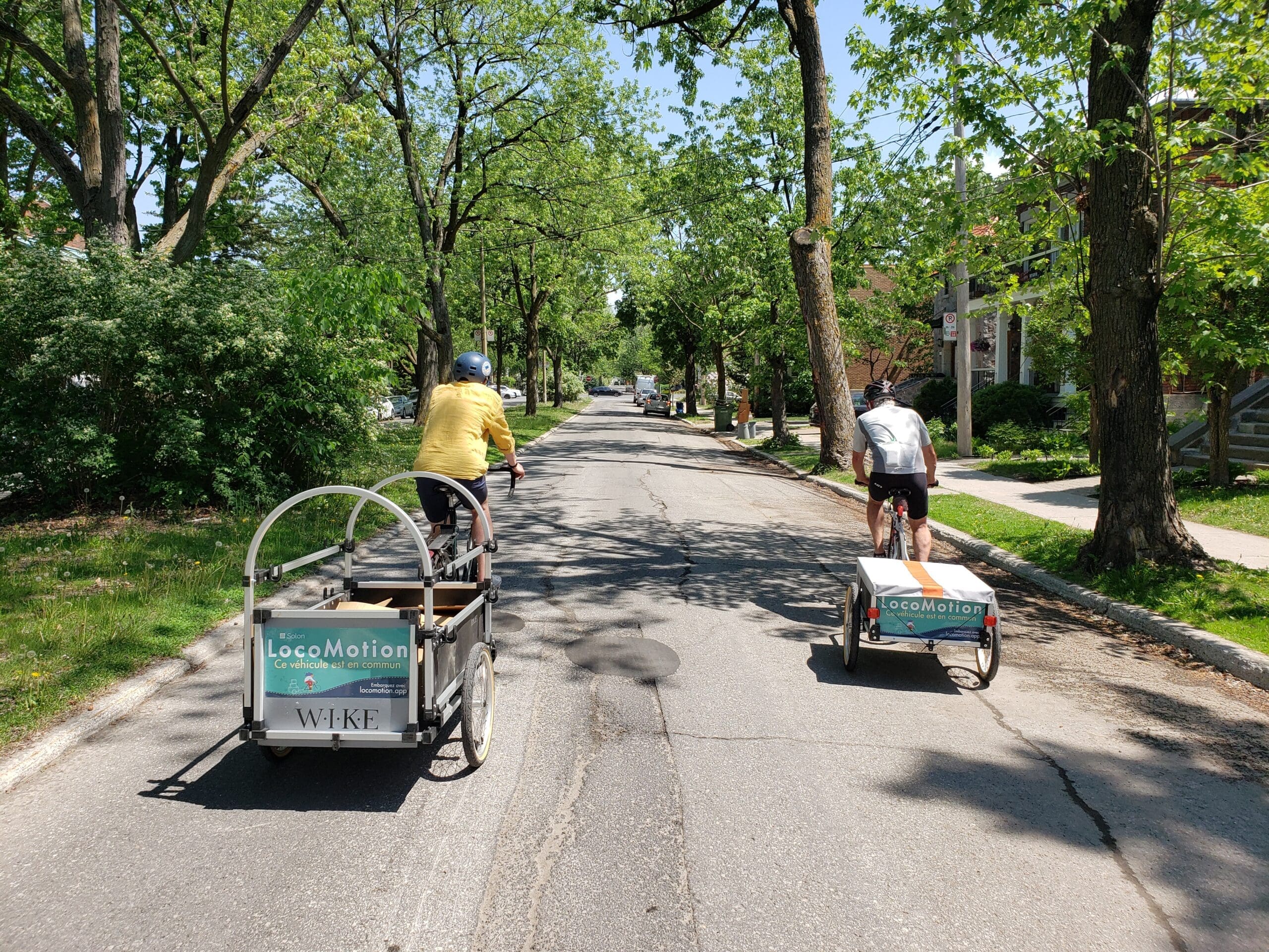 Deux personnes circulent à vélo avec des remorques attachées dans une rue résidentielle bordée d'arbres, mettant en valeur la marque LocoMotion. L'un des cyclistes porte une chemise et un casque jaunes, l'autre un casque blanc. Les arbres et les maisons encadrent magnifiquement la scène - photo prise par LocoMotion.