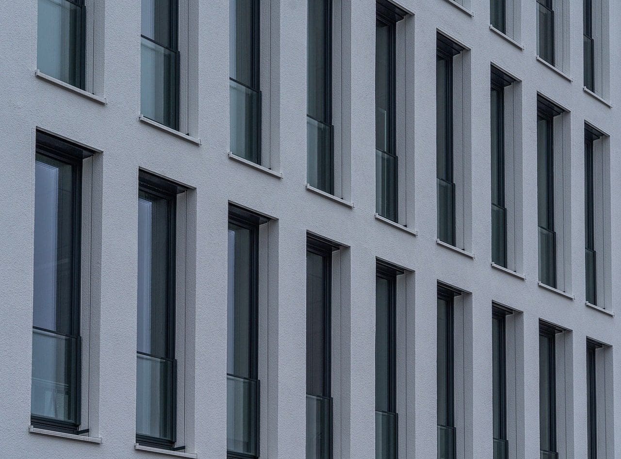 Gros plan sur la façade d'un bâtiment moderne comportant plusieurs rangées et colonnes de grandes fenêtres verticales aux cadres sombres. L'extérieur du bâtiment est lisse et de couleur claire, avec un design minimaliste et symétrique.