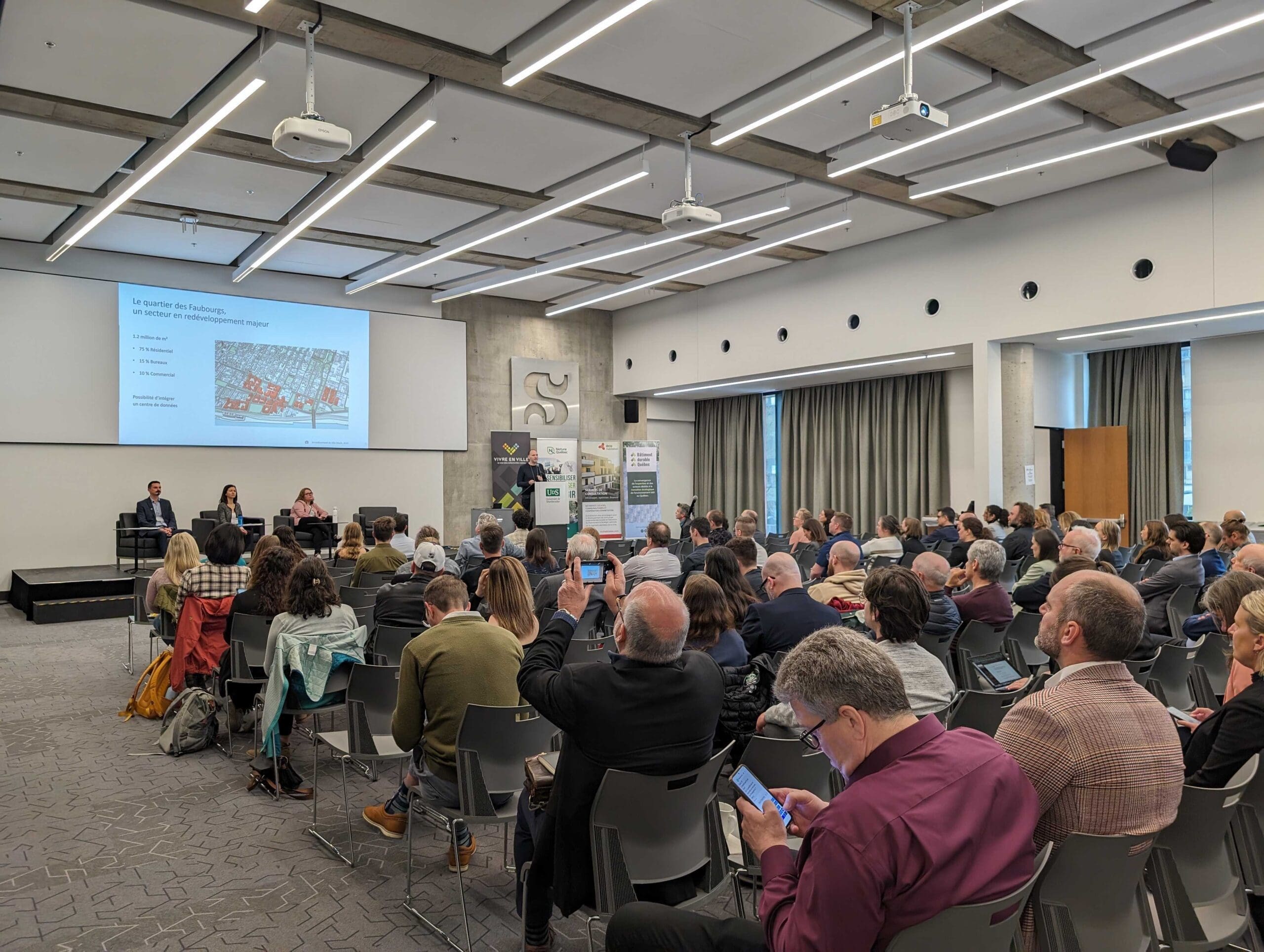 "Un large public est assis dans une salle de conférence et suit attentivement une présentation. Trois orateurs se trouvent à l'avant