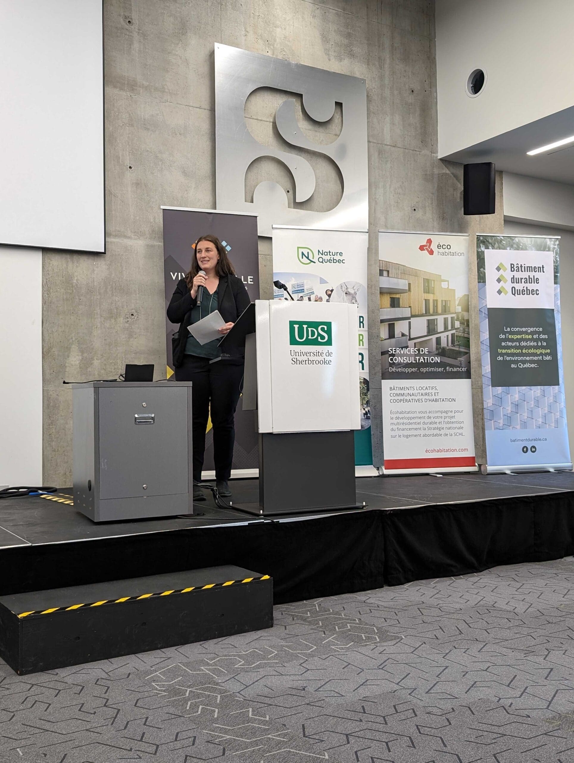 "Une grande salle de conférence bourdonne d'impatience lorsque les participants se rassemblent pour le symposium sur les bâtiments sans carbone. Assis face à un panel de quatre orateurs