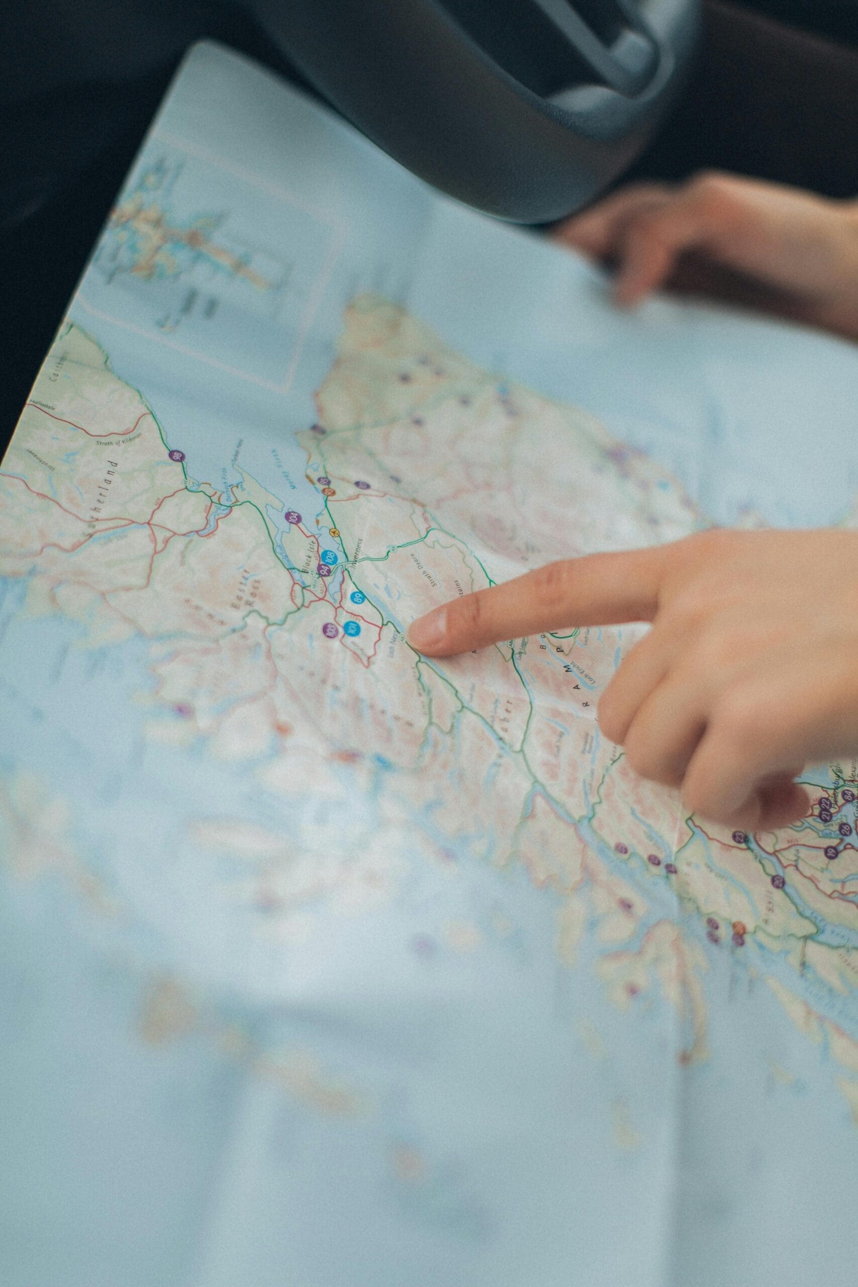 Les mains tiennent et pointent un endroit sur une grande carte imprimée. La carte montre diverses routes et caractéristiques géographiques. La scène suggère la navigation ou la planification d'un voyage. Un volant est partiellement visible à l'arrière-plan.