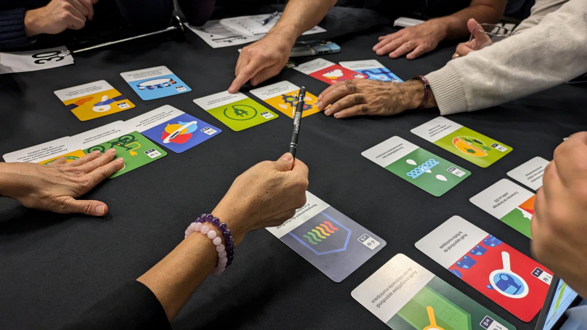 Un groupe réuni autour d'une table est absorbé par le jeu Municipia, chaque membre montrant ou tenant des cartes colorées ornées de graphiques et de symboles intrigants. Ils se concentrent sur la disposition des cartes, qui sont étalées sur une surface sombre, ou en discutent.
