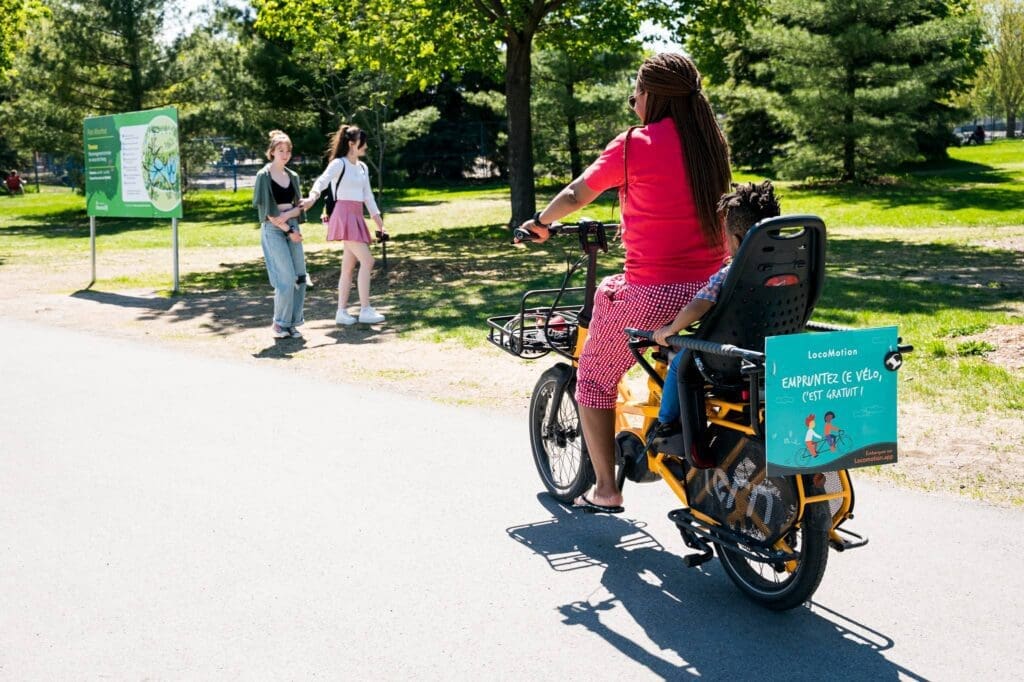 "Une personne aux cheveux tressés roule à vélo sur un chemin avec un enfant dans un siège fixé à l'arrière. Deux personnes marchent à proximité. Le vélo porte un panneau de promotion du tourisme local. Journée ensoleillée avec des arbres et de l'herbe verte."