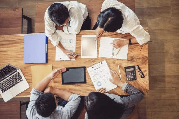 "Quatre personnes sont assises à une table en bois et examinent des documents et des tablettes numériques. Un ordinateur portable