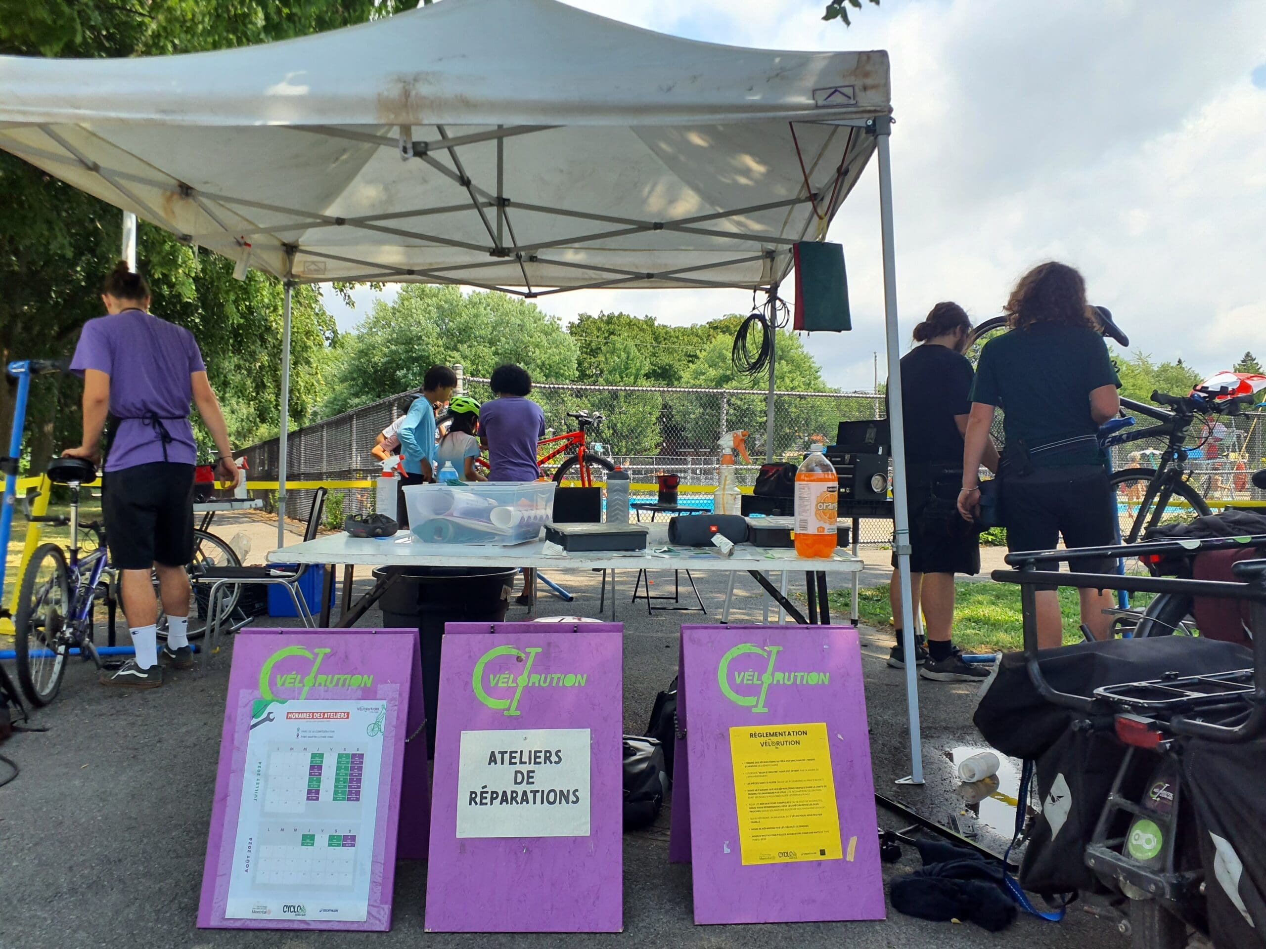 "Des membres du projet Vélorution Couronne réparent des vélos sous une tente blanche lors d'un événement en plein air. Deux panneaux violets affichent en français des informations sur les ateliers de réparation. Des outils sont éparpillés sur la table