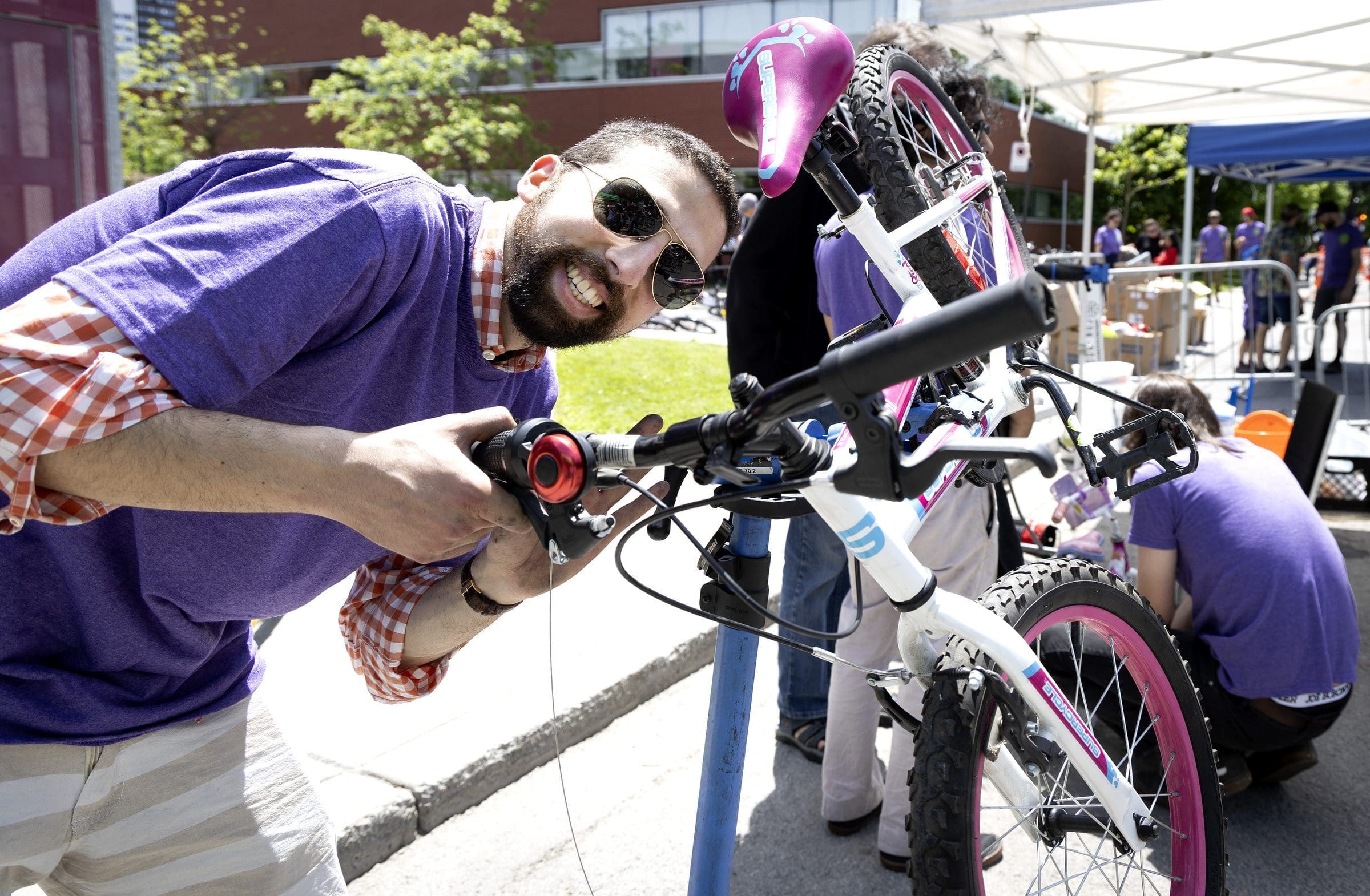 "Un homme souriant portant des lunettes de soleil et une chemise violette répare un vélo rose et blanc dans le cadre du projet Vélorution Couronne. Une autre personne travaille sur un vélo à proximité