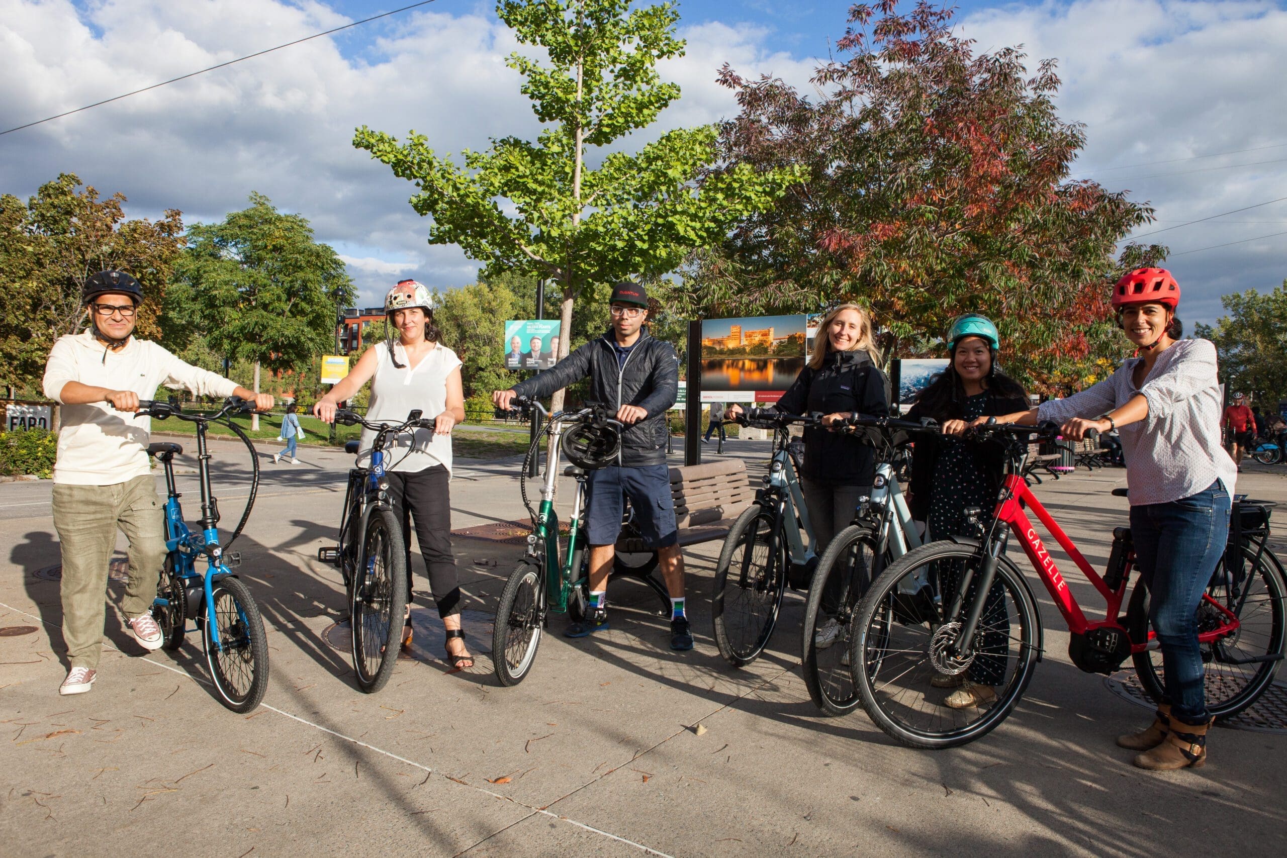 Un groupe de six personnes sourit à l'extérieur, chacun avec un vélo, incarnant l'esprit du projet Vélovolt d'Equiterre. Vêtus de façon décontractée, casque à la main, ils se tiennent debout sur un fond d'arbres, un banc et un ciel partiellement nuageux.
