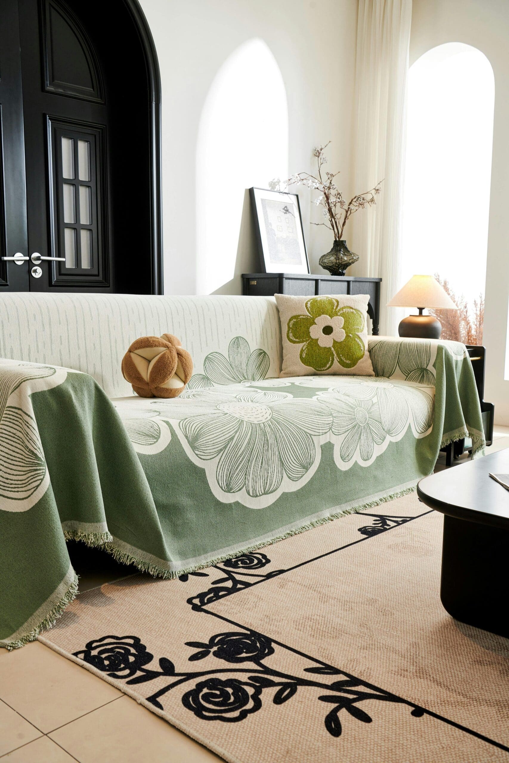 "Un salon élégant présente un canapé drapé d'un jeté vert à motifs floraux. Un coussin orné d'une grande fleur est posé sur le canapé. Une table avec une lampe et des vases se trouve à l'arrière-plan. La pièce est dotée de fenêtres cintrées et d'un tapis à motifs floraux."