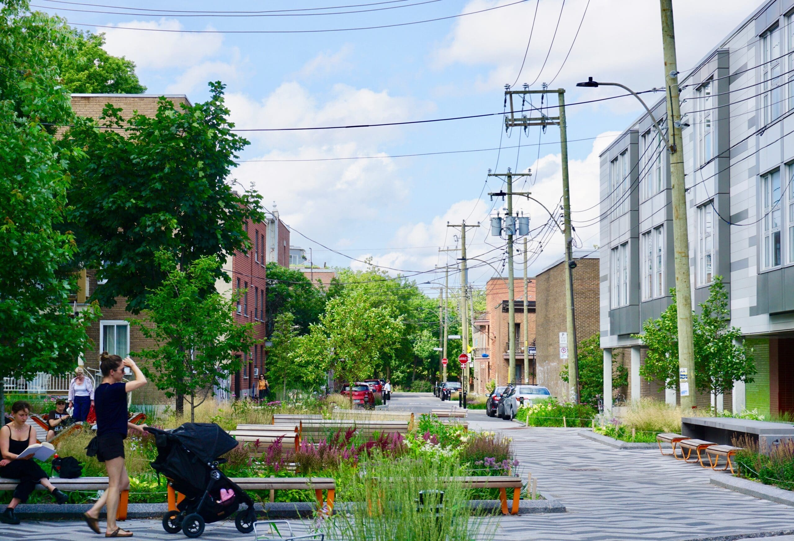 Une scène de rue urbaine animée par Vivre en Ville Montréal montre des gens qui se promènent, y compris une femme poussant une poussette et une autre personne se relaxant sur un banc. La rue, bordée d'arbres et de bâtiments modernes, est traversée par des lignes électriques et le ciel bleu est parsemé de nuages.