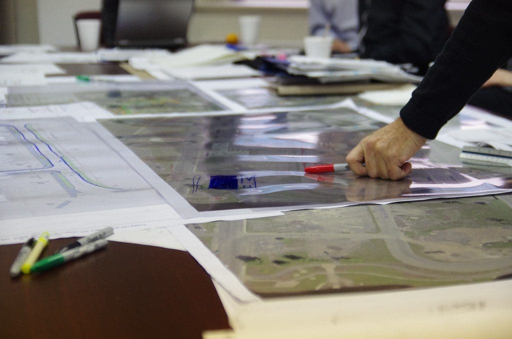 Une personne est penchée sur une table couverte de diagrammes architecturaux et de cartes. Elle tient un marqueur rouge et pointe une section d'une carte. D'autres marqueurs colorés sont éparpillés sur la table.