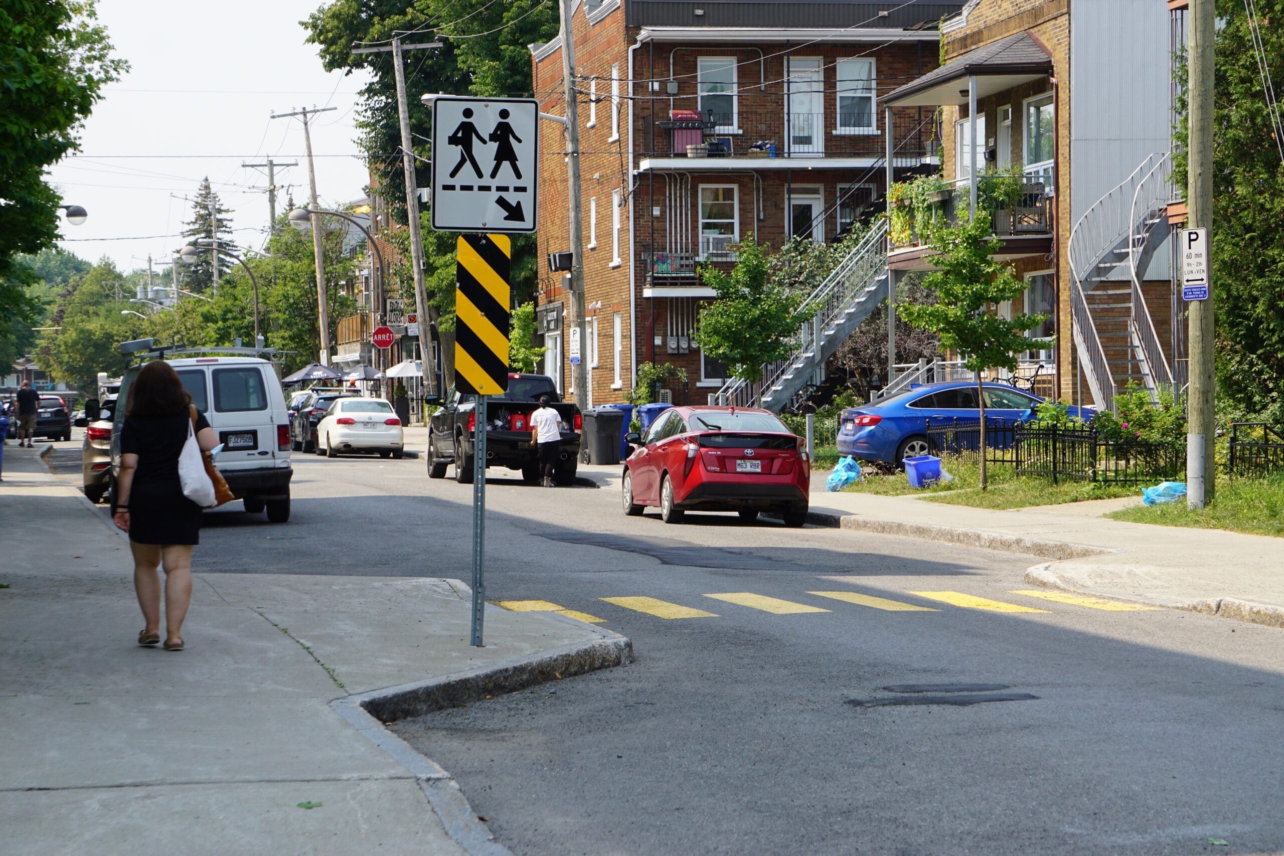 Une femme en robe noire se promène dans une rue de banlieue de Vivre en Ville Montréal. Elle passe devant des voitures garées en rouge, bleu et blanc. Un panneau de passage pour piétons et une route en zigzag sont visibles, avec des arbres en arrière-plan, créant une scène urbaine pittoresque.