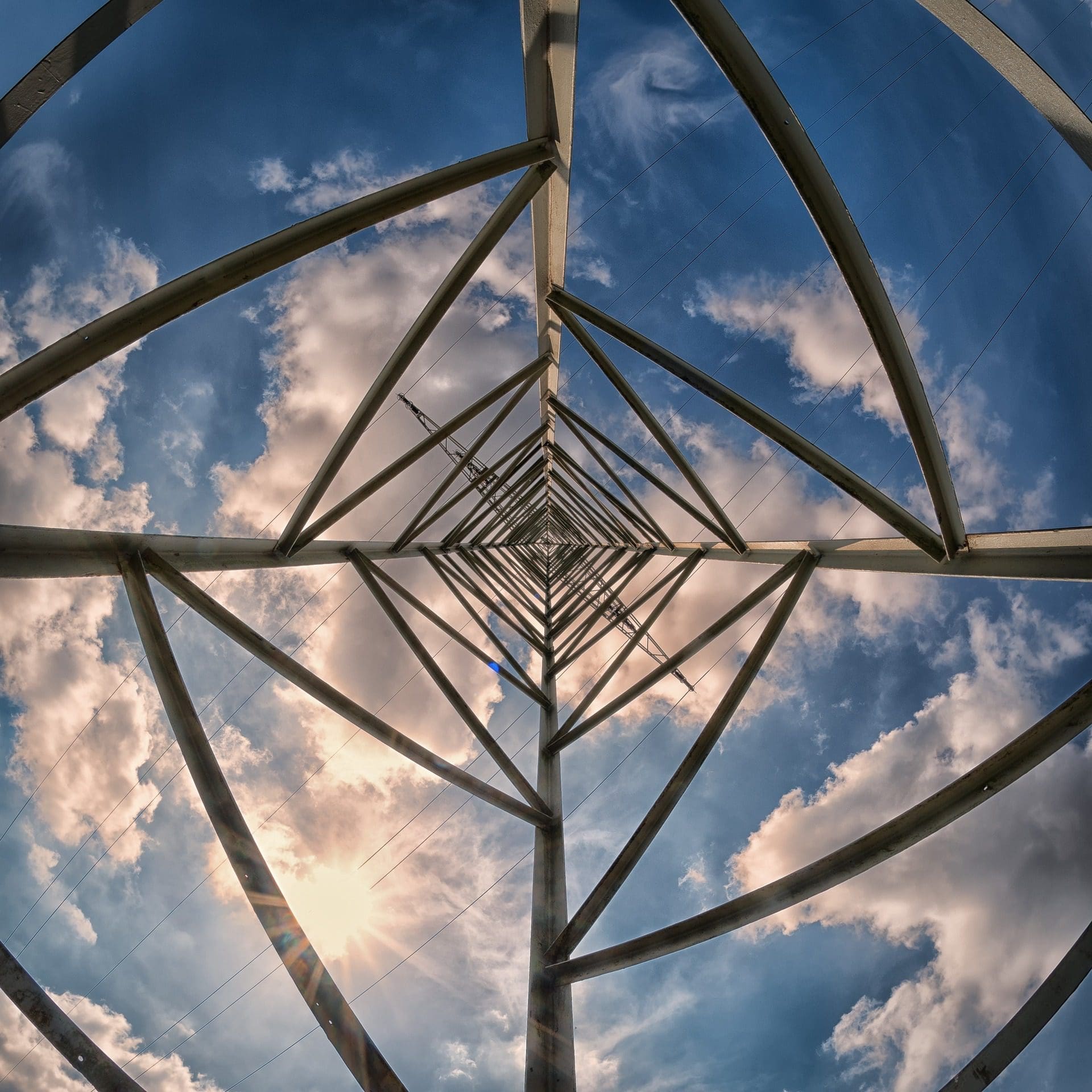 Vue depuis une tour en treillis métallique sur un ciel bleu parsemé de nuages. Le soleil brille à travers la structure, créant un motif de formes géométriques et d'ombres.

