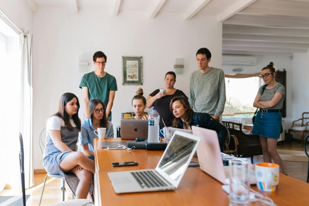 Un groupe de personne observe un ordinateur. Certains sont assis, d'autres debout. L'ambiance est sérieuse, ils travaillent.