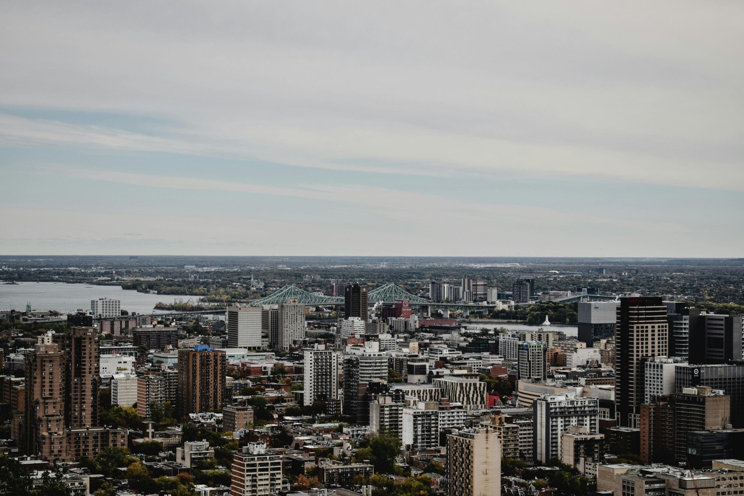 Vue aérienne du paysage urbain de Montréal avec de nombreux gratte-ciel. Un grand pont, rappelant celui photographié par Eva Bronzini, enjambe une rivière à mi-distance sous un ciel partiellement nuageux, avec un horizon de paysages verdoyants en arrière-plan.