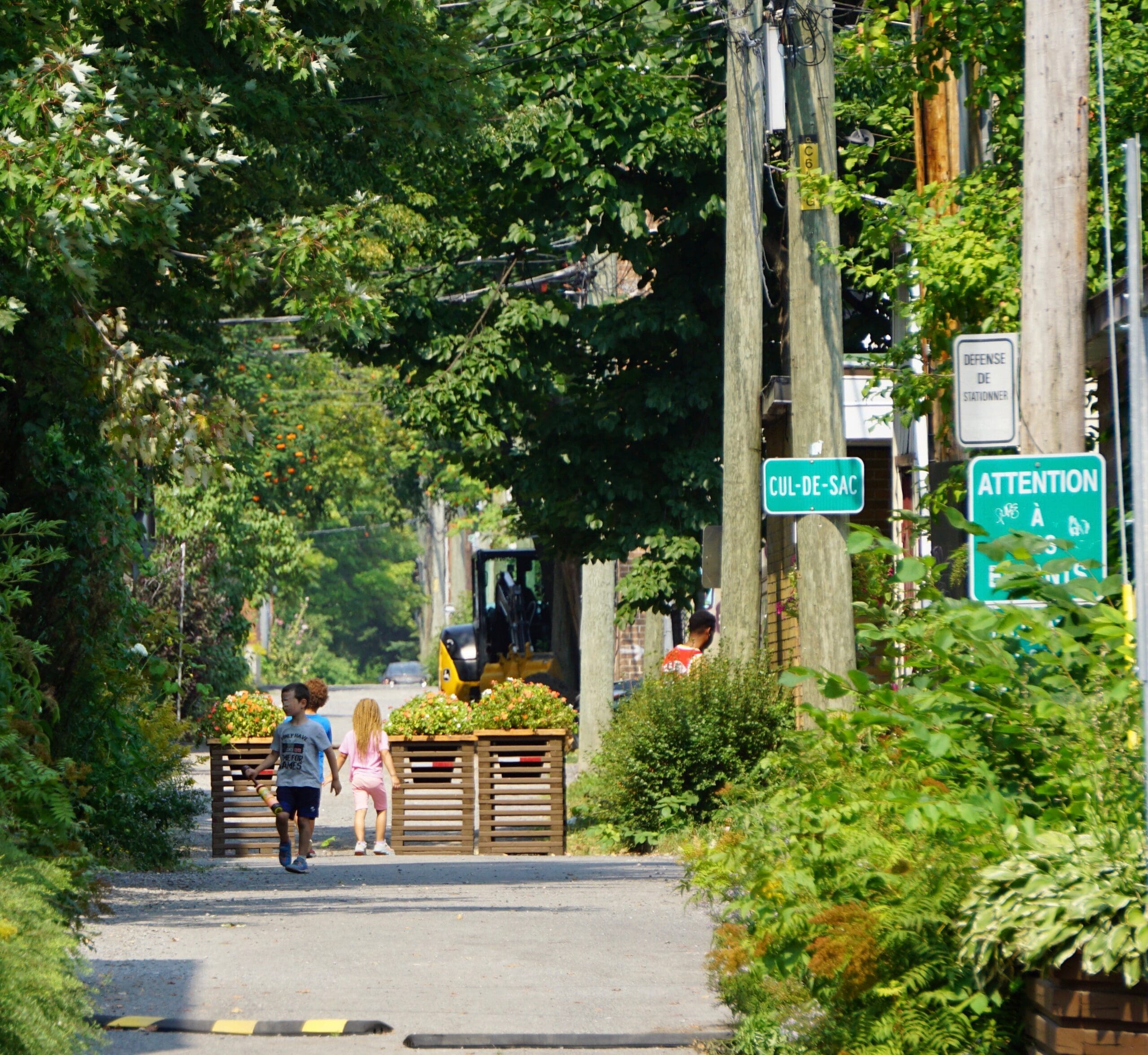 Deux enfants marchent dans une rue étroite, bordée d'arbres, où l'on peut voir des panneaux « cul-de-sac » et « attention ». Un véhicule est garé à l'arrière-plan. Photo prise par Vivre en Ville.