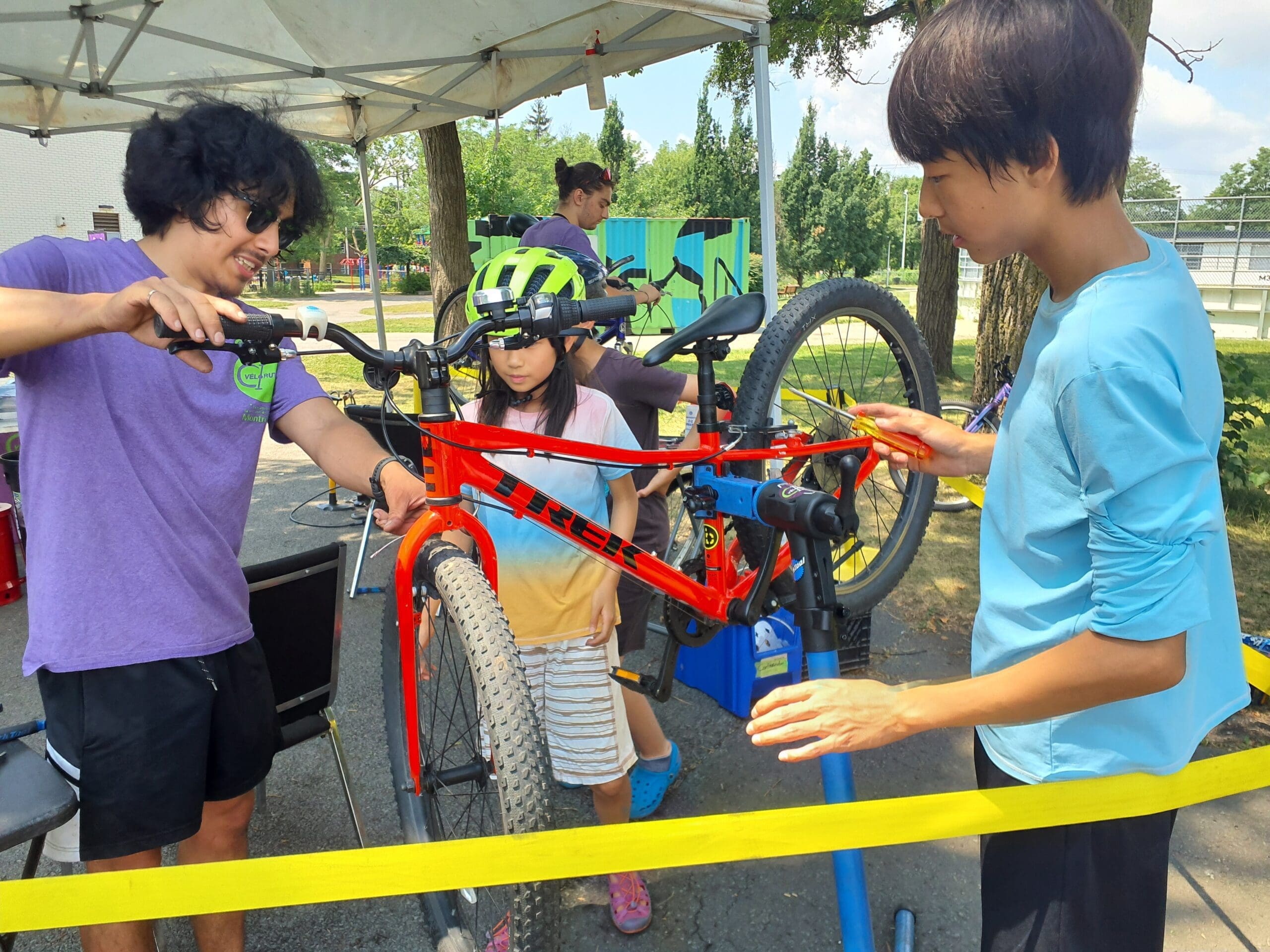 Un enfant avec un casque vert regarde deux personnes réparer un vélo rouge vif sur un stand Vélorution sous une tente. L'une des personnes porte une chemise violette, l'autre une chemise bleue. Des arbres et un parc sont visibles à l'arrière-plan.