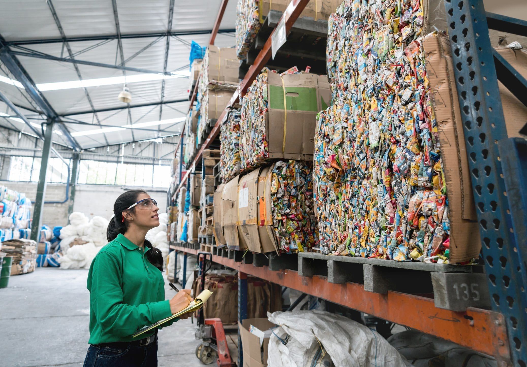 Une personne en chemise verte avec des lunettes de sécurité inspecte des piles de matériaux recyclables compactés dans un entrepôt, en tenant un presse-papiers.