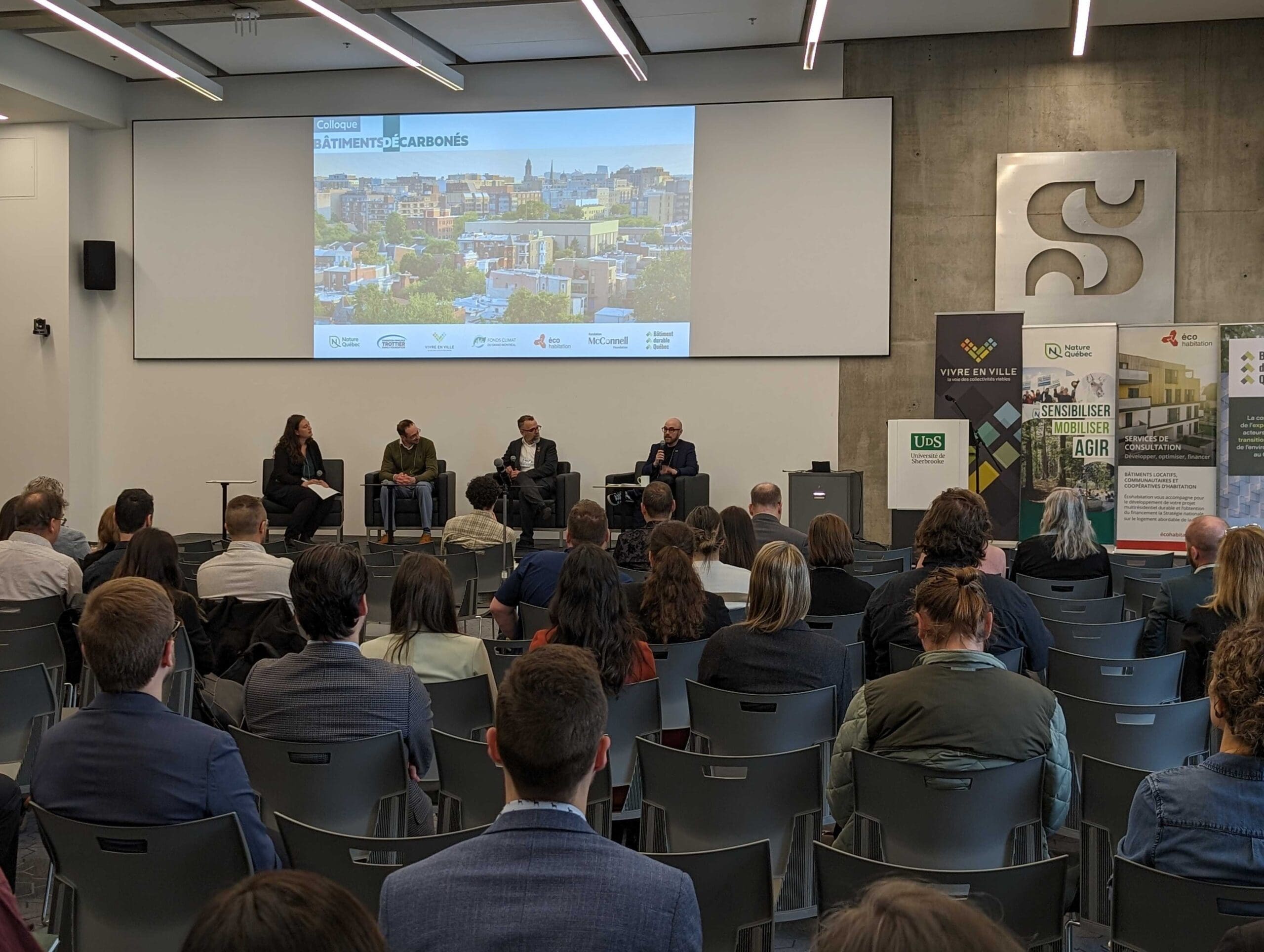 Dans une salle de conférence moderne, quatre panélistes participent à une discussion animée sur les émissions de carbone lors du symposium sur les bâtiments sans carbone (Montréal). Derrière eux, une présentation met en évidence les points clés, tandis que les participants attentifs font face aux orateurs.