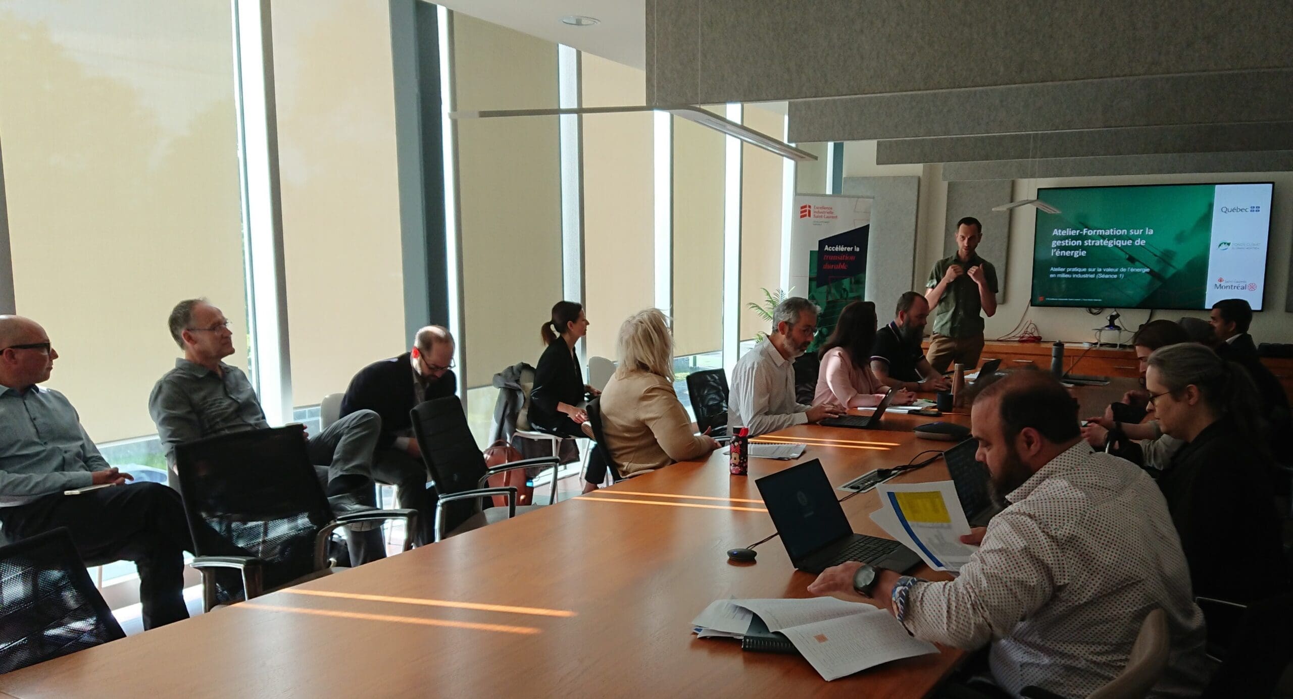 Dans une salle bien éclairée par de grandes fenêtres, des personnes sont assises autour d'une table de conférence pour la réunion sur la boucle énergétique de Saint-Laurent. Une personne se tient près d'un écran affichant une présentation, tandis que d'autres s'activent avec des carnets de notes et des ordinateurs portables ouverts, apportant des idées vibrantes à la discussion.