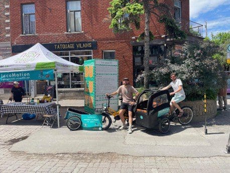 Deux homme en vélo avec une remorque Locomotion devant un stand commercial de locomotion