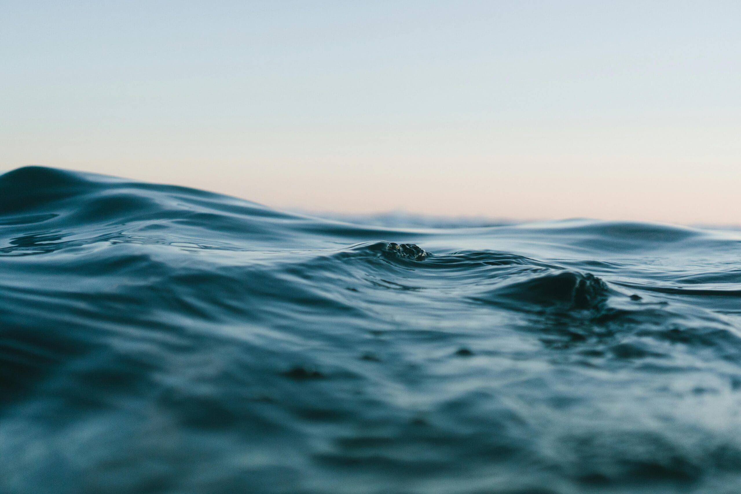 Gros plan sur de douces vagues de l'océan au crépuscule. La surface de l'eau est lisse et ondulée, reflétant de douces teintes bleues et vertes. L'horizon est à peine visible sous un ciel subtil et pâle, créant une atmosphère sereine et paisible.