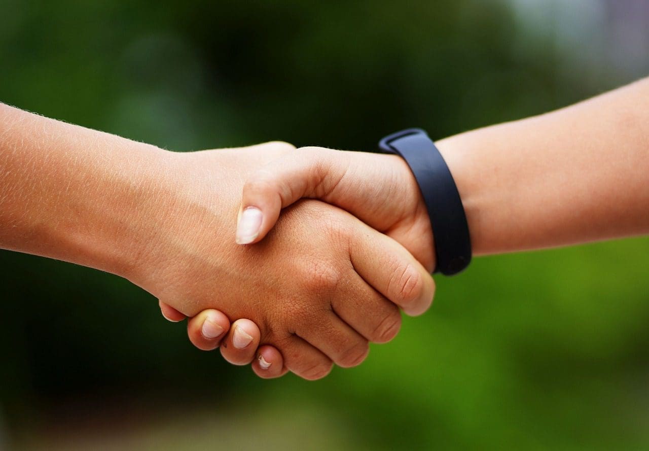 Deux mains se serrent en plein air, l'une d'elles portant un bracelet noir. L'arrière-plan est vert et flou, ce qui suggère un environnement naturel. La poignée de main indique une salutation, un accord ou un partenariat.