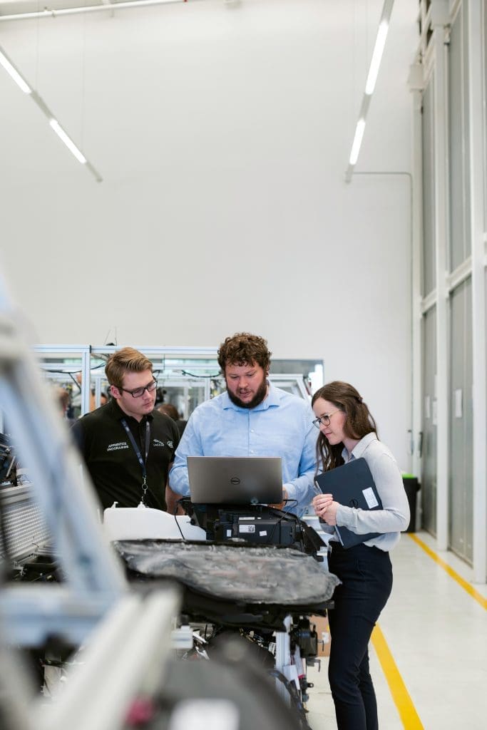 Trois personnes se trouvent dans un environnement industriel lumineux et se concentrent sur un ordinateur portable posé sur un chariot. La personne au centre fait des gestes vers l'écran tandis que les autres écoutent. Ils semblent discuter d'un sujet technique, des machines étant visibles en arrière-plan.