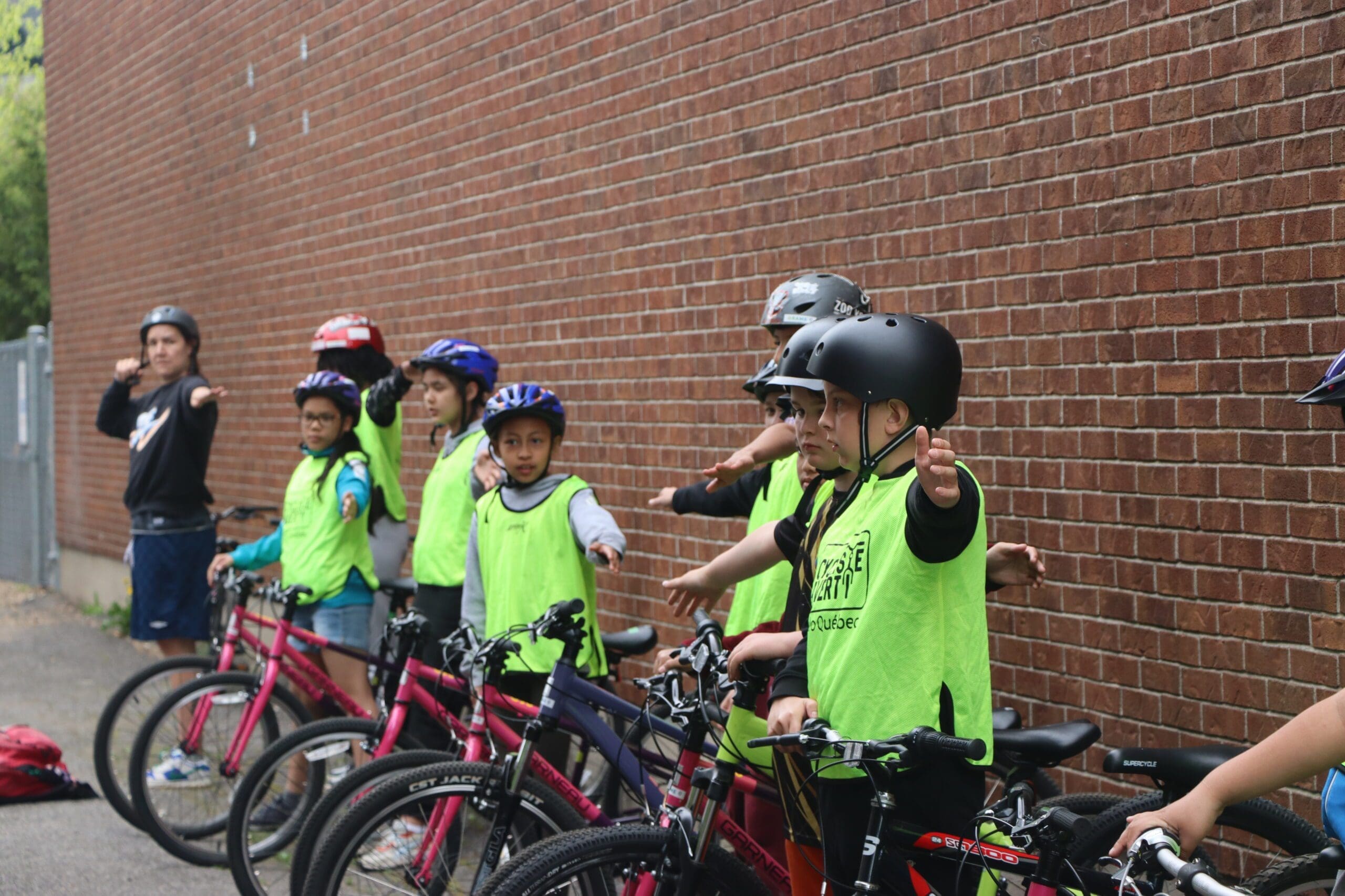 Des enfants portant des casques et des gilets de sécurité verts se tiennent près de leurs vélos le long d'un mur de briques, participant au programme Cycliste Averti de Vélo Québec. Les bras tendus, ils font des signaux sous la direction d'un adulte instructeur.