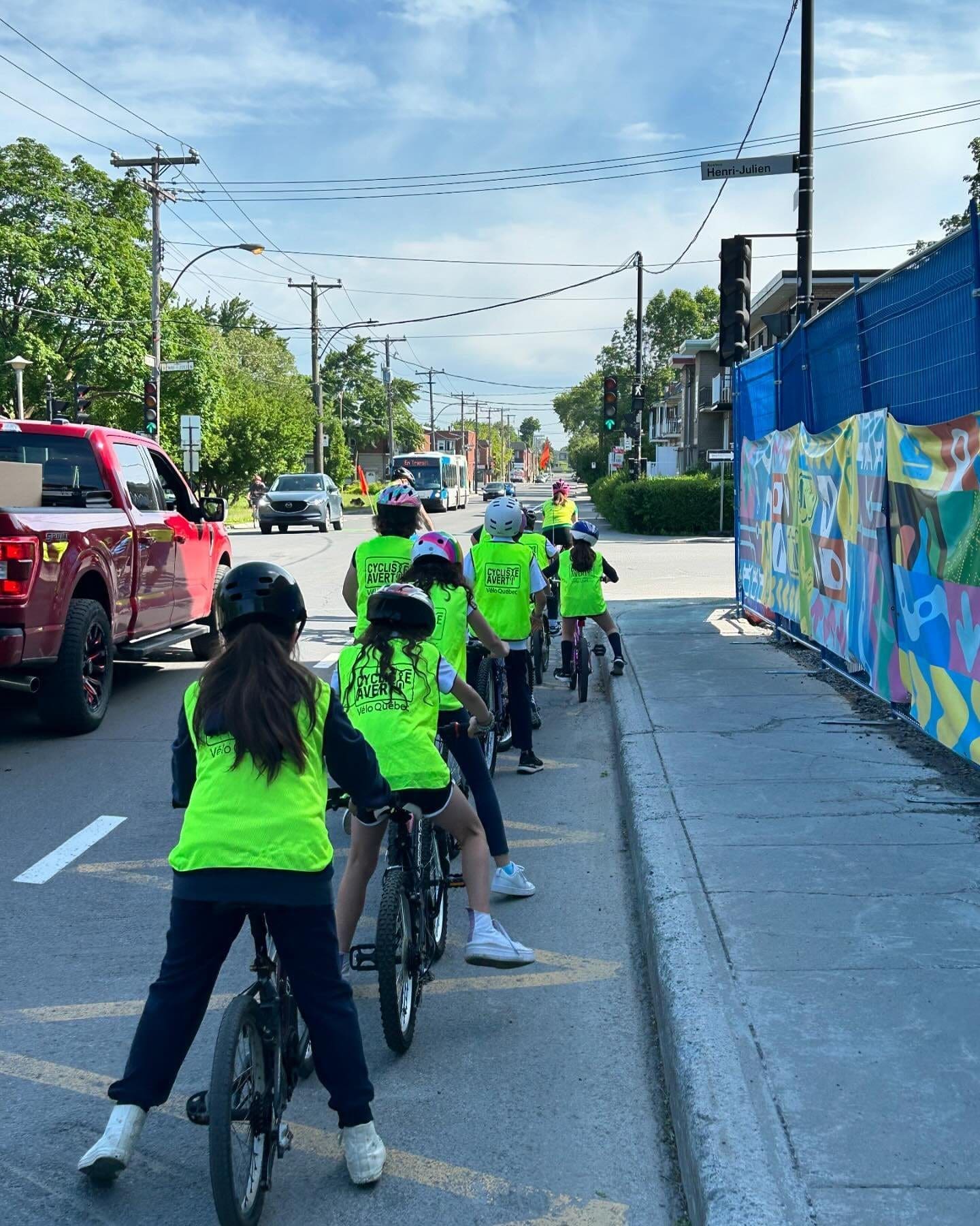 Un groupe d'enfants vêtus de gilets jaune vif, faisant partie du programme Cycliste Averti de Vélo Québec, roule à vélo en file indienne dans une rue de la ville. Ils passent devant un trottoir et une peinture murale colorée, avec des voitures et des arbres en arrière-plan, sous un ciel partiellement nuageux.