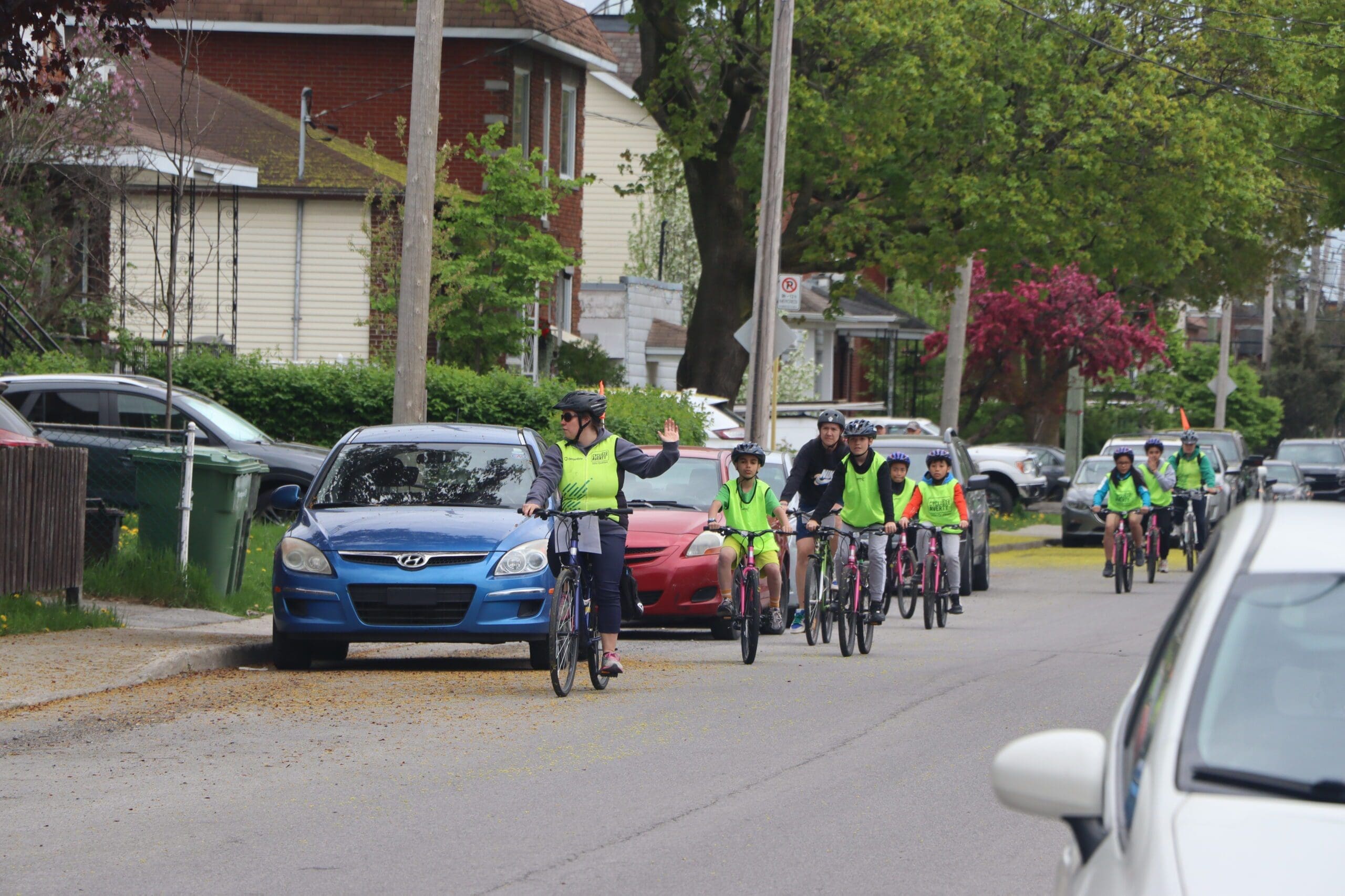 Un groupe de cyclistes participant au programme Cycliste Averti de Vélo Québec roule en file indienne dans une rue de banlieue, vêtus de gilets de sécurité vert vif. La femme à l'avant fait un signe du bras tandis que les arbres, les voitures garées et les maisons se tiennent tranquillement à l'arrière-plan.