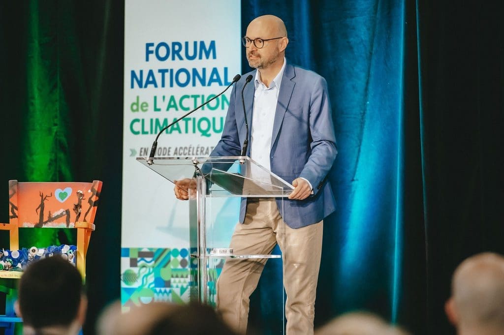 Une personne vêtue d'un blazer bleu et d'un pantalon beige s'adresse à un podium muni d'un microphone lors de l'événement FNAC2024 du RNCREQ. En arrière-plan, on peut voir un panneau indiquant Forum National de l'Action Climatique. Une chaise colorée et une petite table sont visibles à gauche.