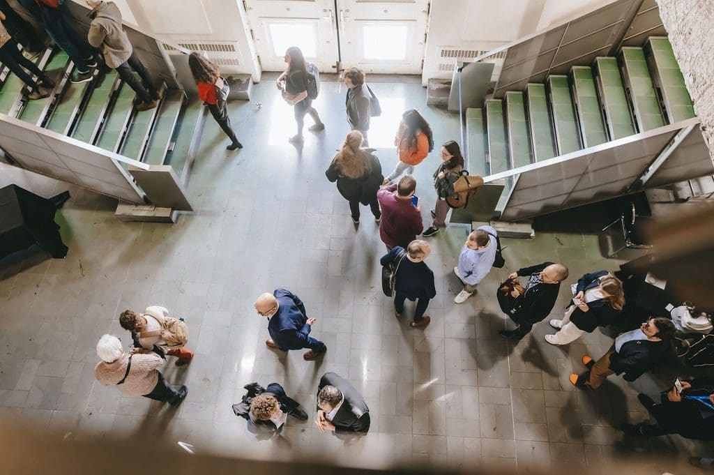 Vue aérienne de l'événement FNAC2024 par le RNCREQ montrant des personnes rassemblées dans un couloir. Un groupe se tient près d'un escalier, tandis que d'autres discutent. Le sol est carrelé et la lumière naturelle passe à travers de grandes fenêtres, ajoutant de la chaleur à l'atmosphère animée.