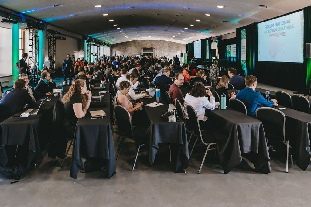 Une grande salle de conférence remplie de personnes assises à des tables, faisant face à une scène où le Forum National sur l'Action Climatique orne l'écran de présentation. Cet événement FNAC2024 du RNCREQ est bien éclairé et les participants sont activement engagés dans les discussions.