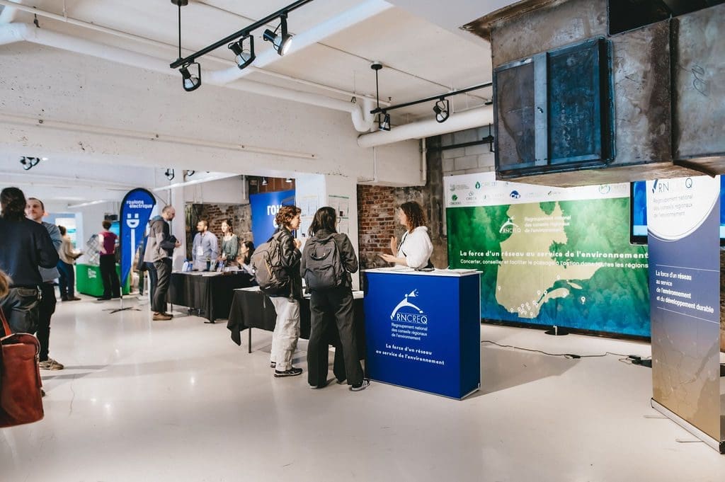 Les gens sont rassemblés au FNAC2024, un événement organisé par le RNCREQ, qui présente divers stands d'information sur des sujets environnementaux. Les tables sont garnies de brochures et de bannières, et les participants participent à des discussions. L'endroit est bien éclairé et présente une ambiance industrielle moderne.