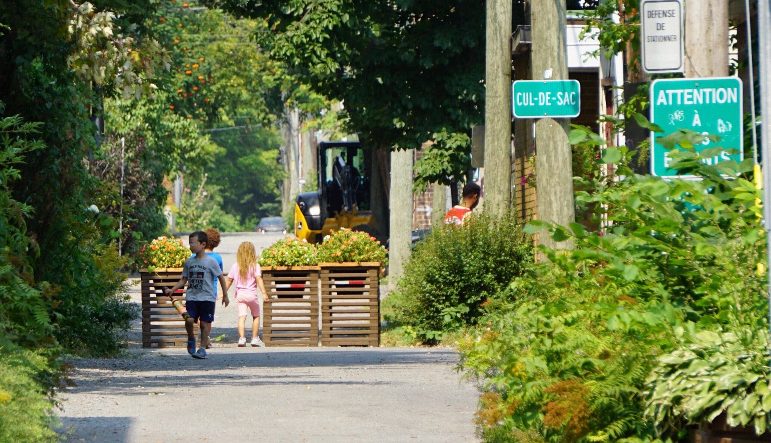 Deux enfants marchent dans une rue étroite, bordée d'arbres, où l'on peut voir des panneaux « cul-de-sac » et « attention ». Un véhicule est garé à l'arrière-plan.