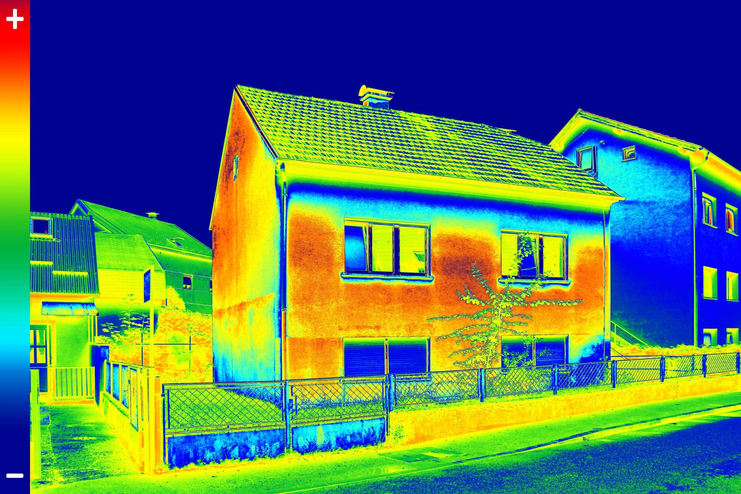 Une image thermique d'une rue résidentielle montre une maison à deux étages avec des variations de température. La maison est principalement rouge et orange, ce qui indique des zones plus chaudes, avec des teintes bleues et vertes plus froides sur le toit et les bâtiments voisins.