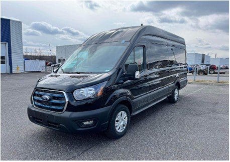 Fourgonnette Ford Transit noire de La centrale roule ! garée sur une surface asphaltée dans un parking. La camionnette a un toit haut et des vitres teintées. Le ciel est partiellement nuageux et plusieurs bâtiments et véhicules sont visibles à l'arrière-plan.