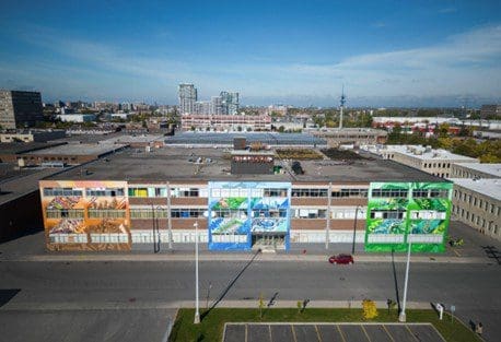 Une vue aérienne de la Coop Centrale Agricole, un grand bâtiment multicolore orné de peintures murales vibrantes. Entouré de structures industrielles, il se détache au milieu de quelques véhicules garés et d'un terrain vague au premier plan. Le ciel clair est parsemé de nuages épars.