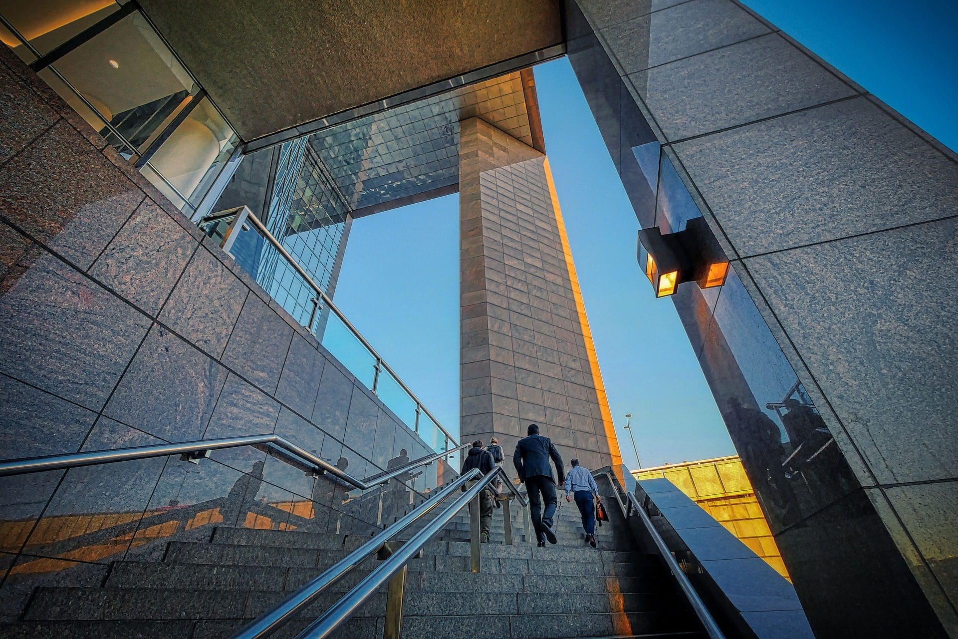 Des personnes montent un large escalier flanqué d'une architecture moderne et de surfaces vitrées réfléchissantes. La structure est éclairée par la lueur chaude du soleil couchant, qui projette des ombres et met en valeur le design angulaire dans un ciel bleu clair.