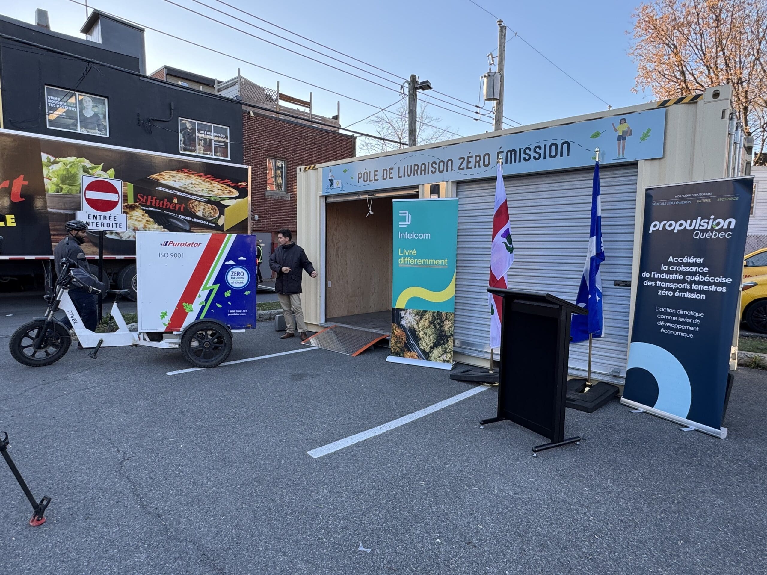 Sur le site de l'événement en plein air, un vélo cargo avec une boîte de livraison affiche fièrement des bannières pour la livraison zéro émission en français par Propulsion Québec. Un podium pour les conférenciers complète la scène, tandis qu'une remorque avec une image vibrante de nourriture se trouve à l'arrière-plan du parking.