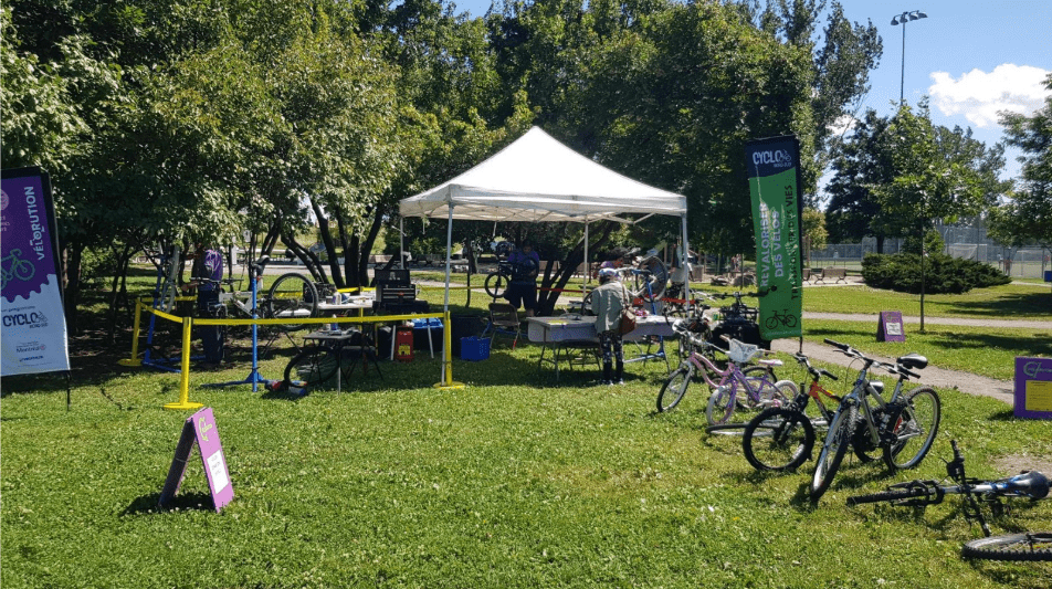 Une petite station de réparation de vélos en plein air pour l'événement MobiliSENS se compose d'une tente blanche dans un parc herbeux. Plusieurs vélos sont installés autour de la tente, avec des outils de réparation à portée de main. Des arbres et un ciel bleu offrent une toile de fond sereine, tandis que des panneaux affichant les logos de l'événement sont placés à proximité.