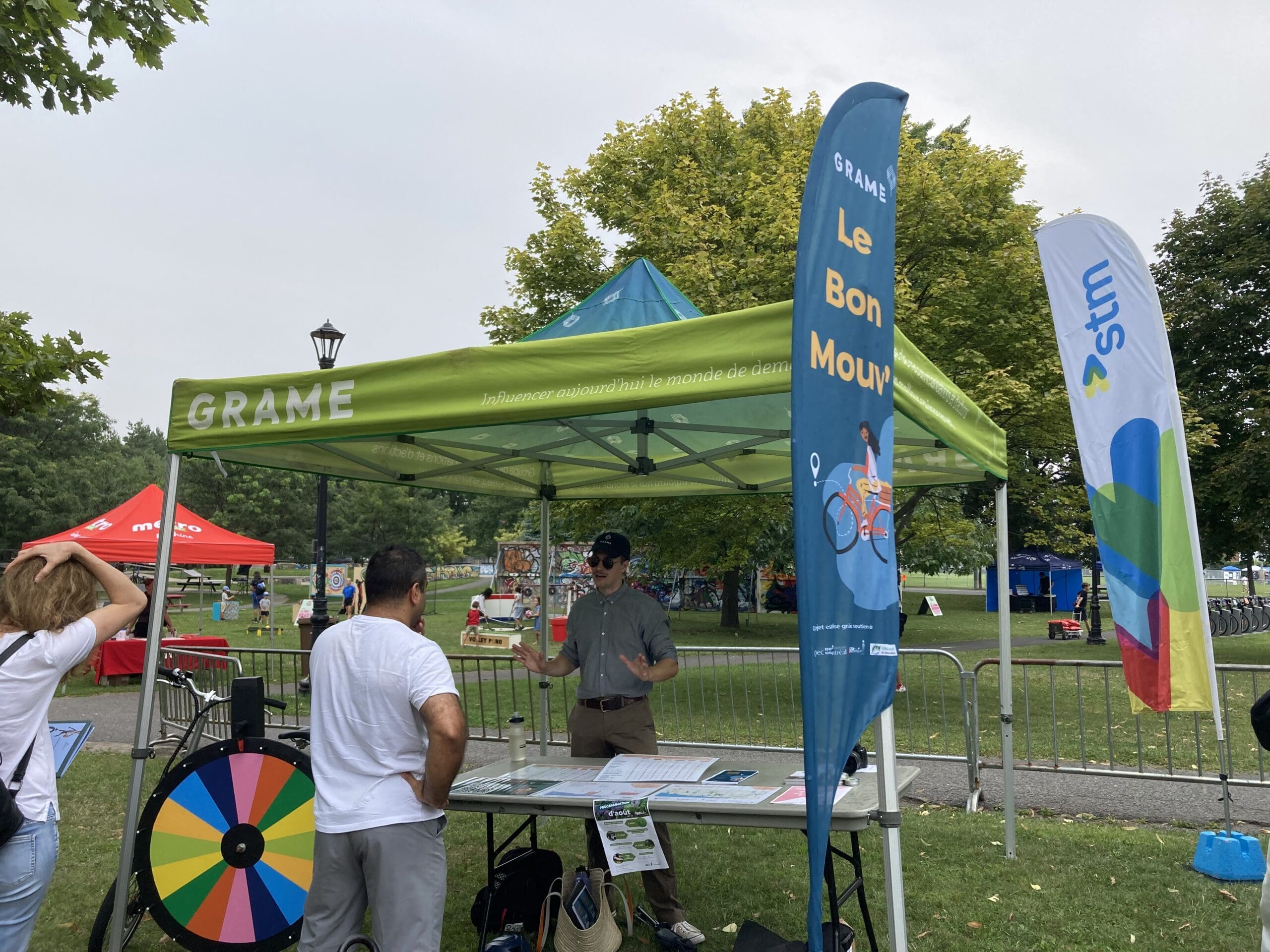 Un petit stand extérieur avec un auvent vert portant le nom de GRAME Le Bon Mouv lors d'un événement. Il y a des bannières, une roue colorée sur un support et une personne en chemise bleue qui discute avec les visiteurs. Des arbres luxuriants et d'autres stands créent une toile de fond vivante.