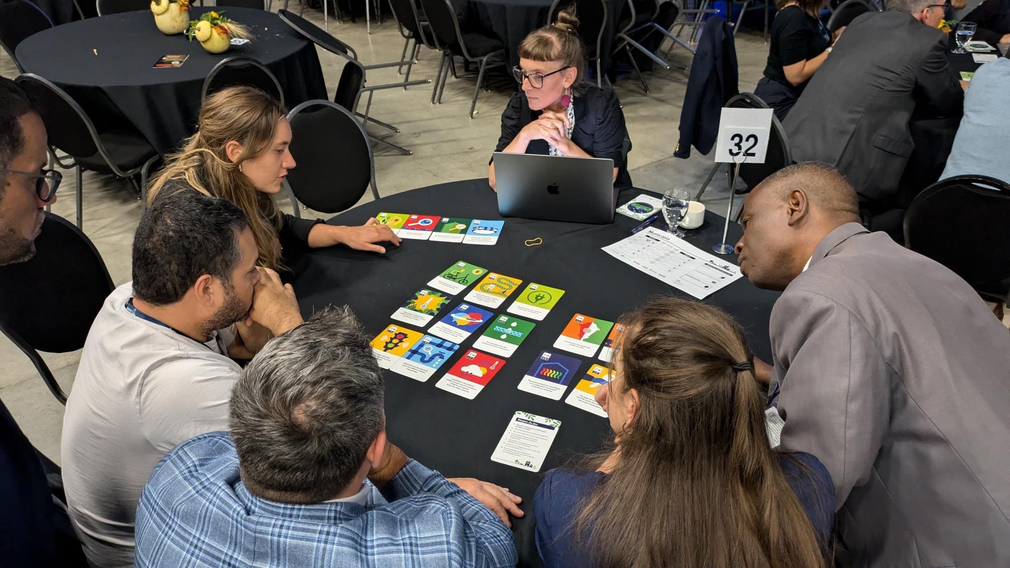 Un groupe de sept personnes est assis autour d'une table, jouant au jeu Municipia d'IVÉO au SIIViM Drummondville avec des cartes colorées étalées devant eux. Une personne utilise un ordinateur portable. L'ambiance est à la collaboration et à la concentration, avec une carte de numéro de table indiquant 32 bien en vue.