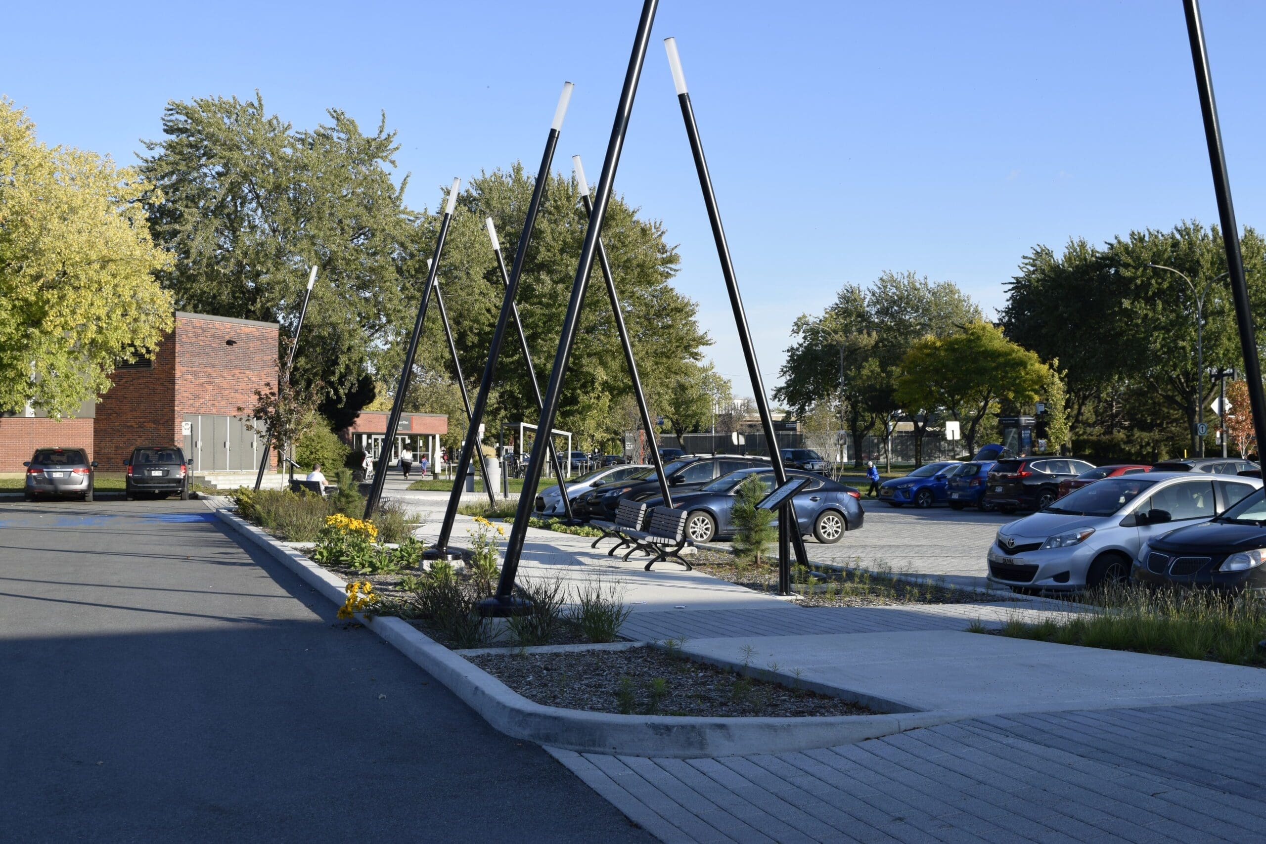 Un parking avec plusieurs voitures est bordé de hautes sculptures noires. À gauche, il y a un bâtiment en briques et, à l'arrière-plan, des arbres aux feuilles vertes et jaunes. Le ciel est clair et bleu.