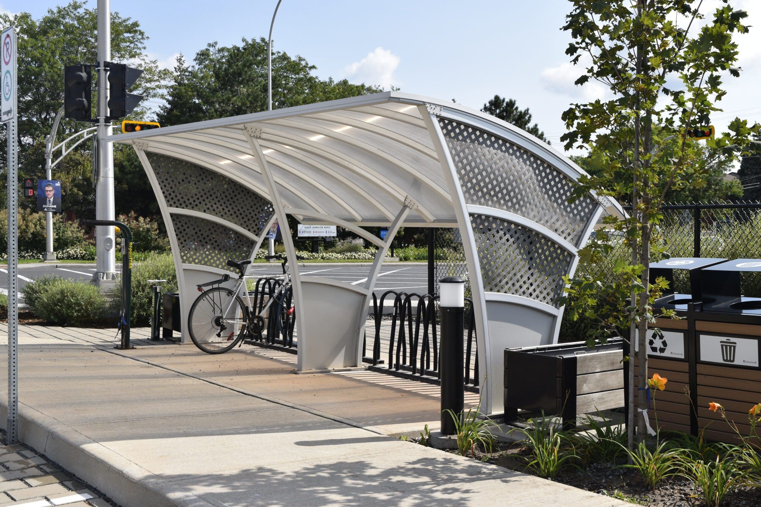 Un abri à vélos moderne, blanc, avec un toit arqué et des panneaux perforés. À l'intérieur, plusieurs vélos sont garés. L'abri est entouré d'un jardin bien entretenu et une station de recyclage est visible à proximité. Une route et des feux de signalisation se trouvent à l'arrière-plan.