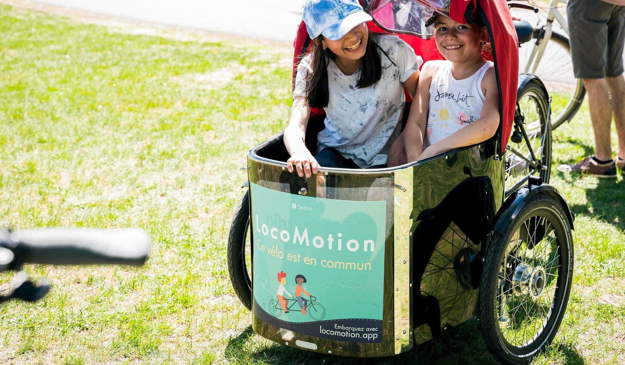 Deux enfants sont assis dans un vélo cargo par une journée ensoleillée. Ils sourient et portent des casquettes. Le vélo porte un panneau indiquant LocoMotion ainsi qu'un texte en français. Des personnes et un vélo sont visibles à l'arrière-plan sur un terrain herbeux.