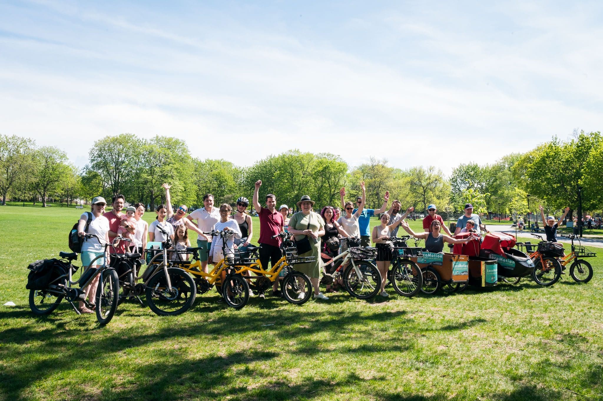 Un groupe de personnes pose avec enthousiasme sur des vélos cargo LocoMotion dans un parc ensoleillé, crédit Audrey McMahon. Certains lèvent les bras en signe d'excitation sur fond d'herbe verte et d'arbres sous un ciel bleu limpide.