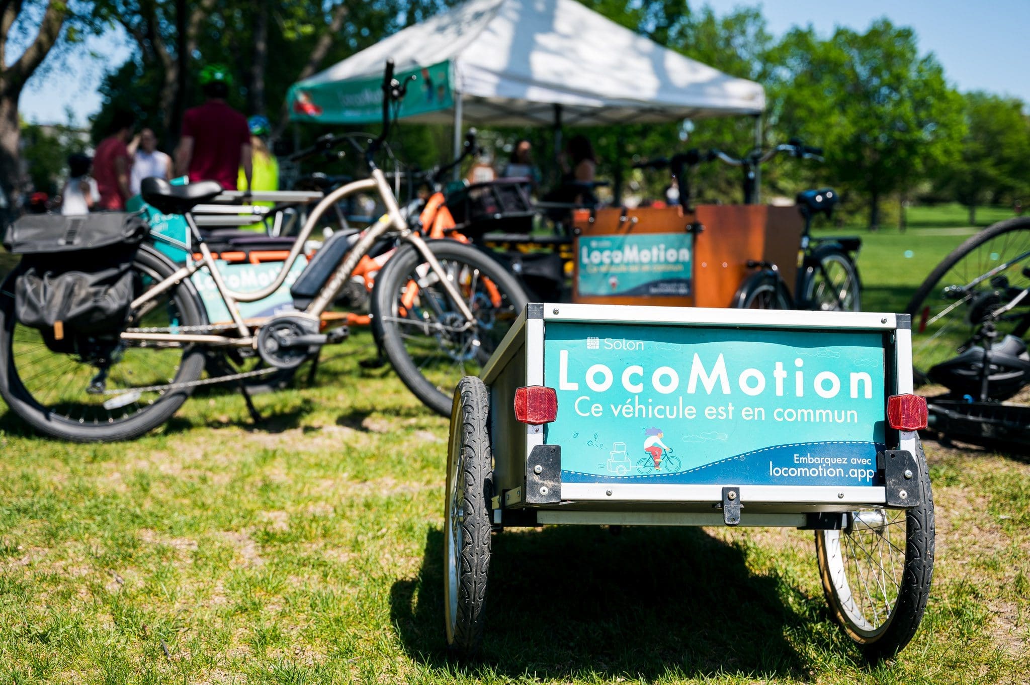 Des vélos et une remorque avec un panneau LocoMotion sont posés sur l'herbe près d'une tente blanche. Des gens se rassemblent à l'arrière-plan par une journée ensoleillée, les arbres étant visibles derrière eux. Crédit : Audrey McMahon