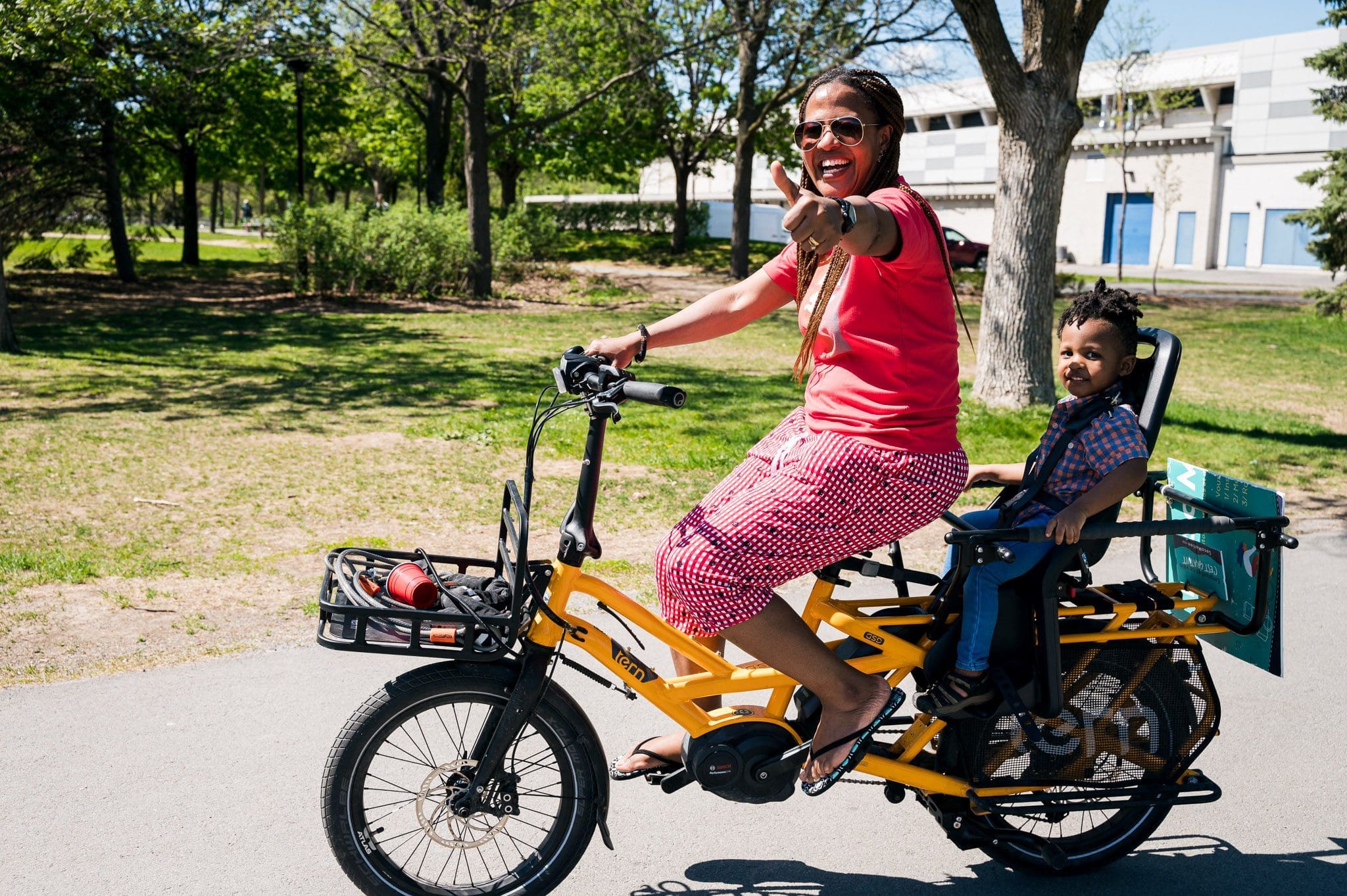 Une femme portant des lunettes de soleil et un tee-shirt rouge conduit un vélo électrique jaune dans le parc, en souriant et en montrant du doigt la direction à suivre. Un jeune enfant est assis sur le siège arrière, s'accrochant au guidon, tous deux profitant de la journée ensoleillée avec des arbres et un bâtiment derrière eux. Audrey McMahon a capturé cette joie.